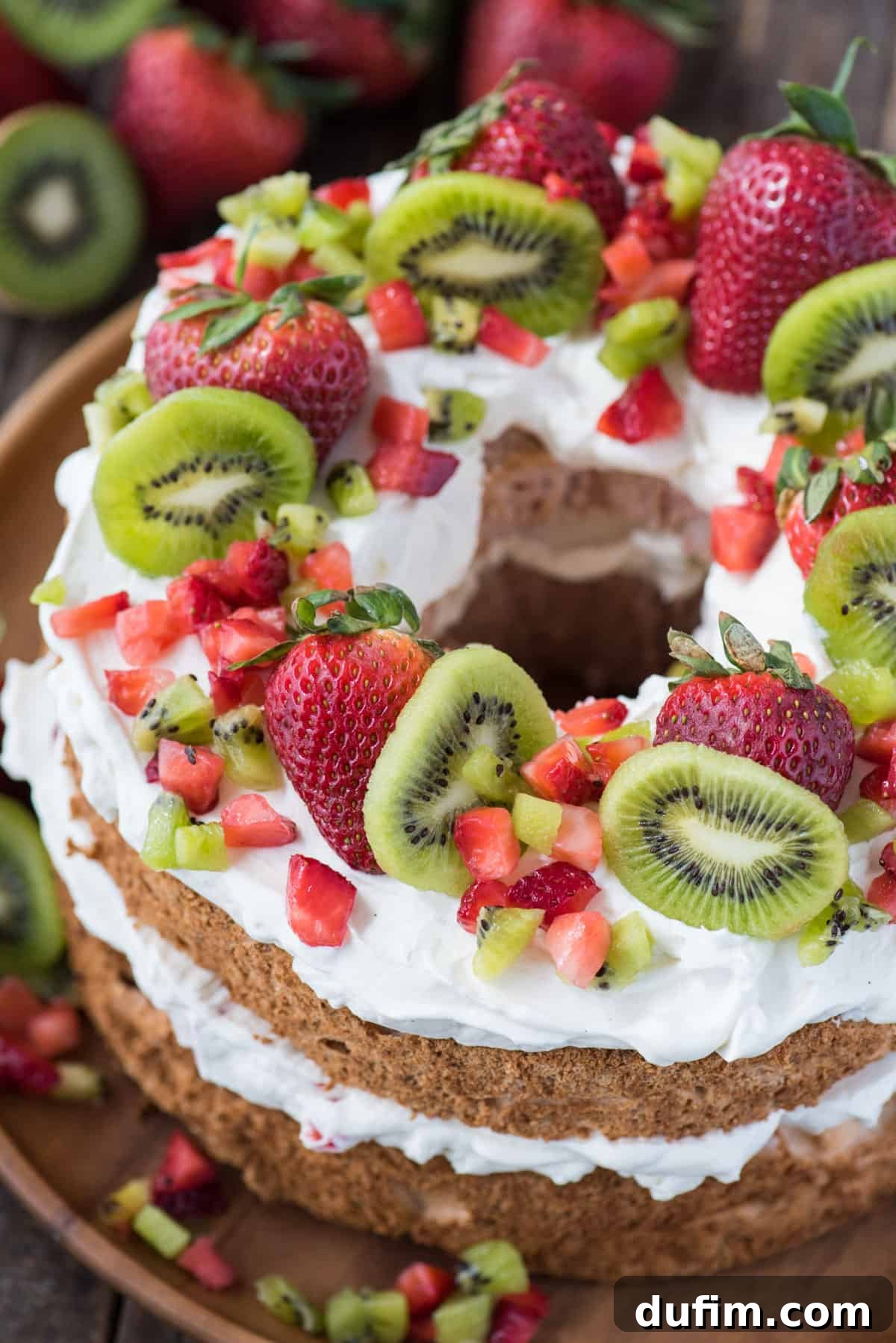Heavenly Strawberry Cloud Cake 5 strawberry angel food cake topped with whipped cream, whole strawberries, chopped strawberries and kiwi on wooden serving tray