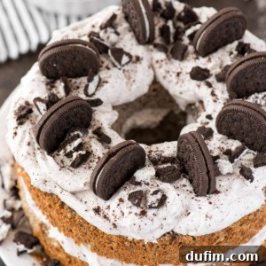 A round oreo angel food cake with white frosting, topped with whole and crushed chocolate sandwich cookies, and more cookies pressed between the layers, served on a white plate.