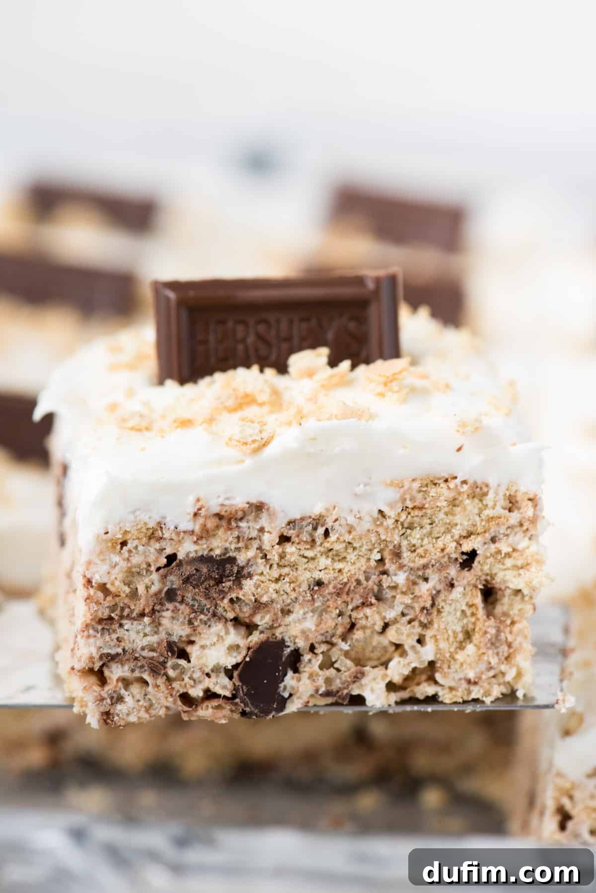 No-Bake S'mores Rice Krispie Treats 7 A single S'mores Rice Krispie treat, featuring a generous dollop of marshmallow frosting and a piece of Hershey's bar, elegantly presented on a metal serving utensil.