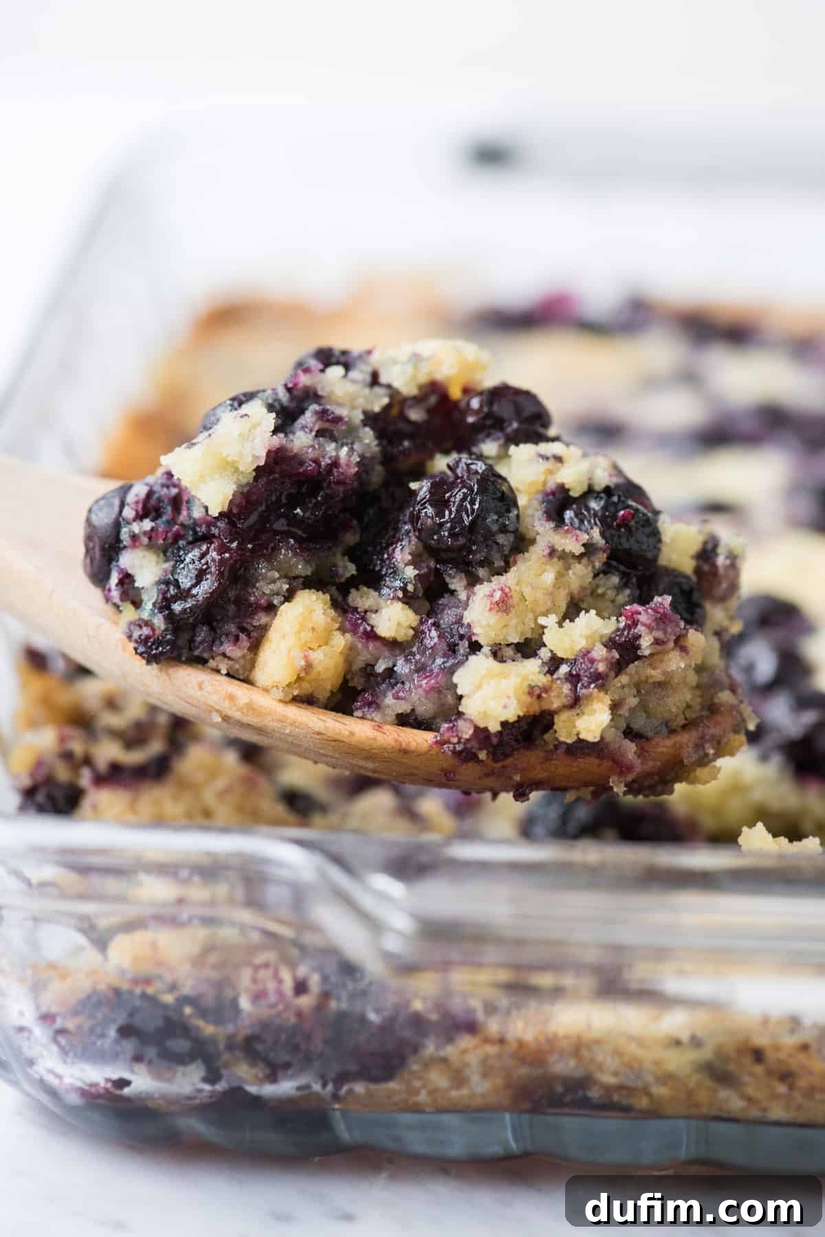 Zesty Lemon Blueberry Bake 2 Lemon Blueberry Dump Cake in a glass 9x13 inch pan, with a wooden spoon scooping out a generous portion, showcasing the golden crust and fruity interior.