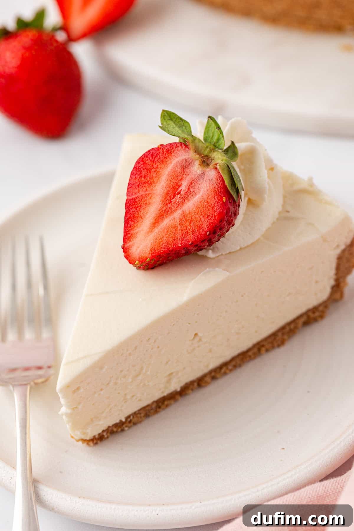 Whip Up a Philadelphia No Bake Cheesecake 2 A beautifully presented slice of creamy Philadelphia no-bake cheesecake featuring a golden graham cracker crust, elegantly topped with swirls of homemade whipped cream and a fresh half-strawberry. It rests on a pristine white plate, with a fork gently placed beside it, hinting at its inviting texture and readiness to be savored.