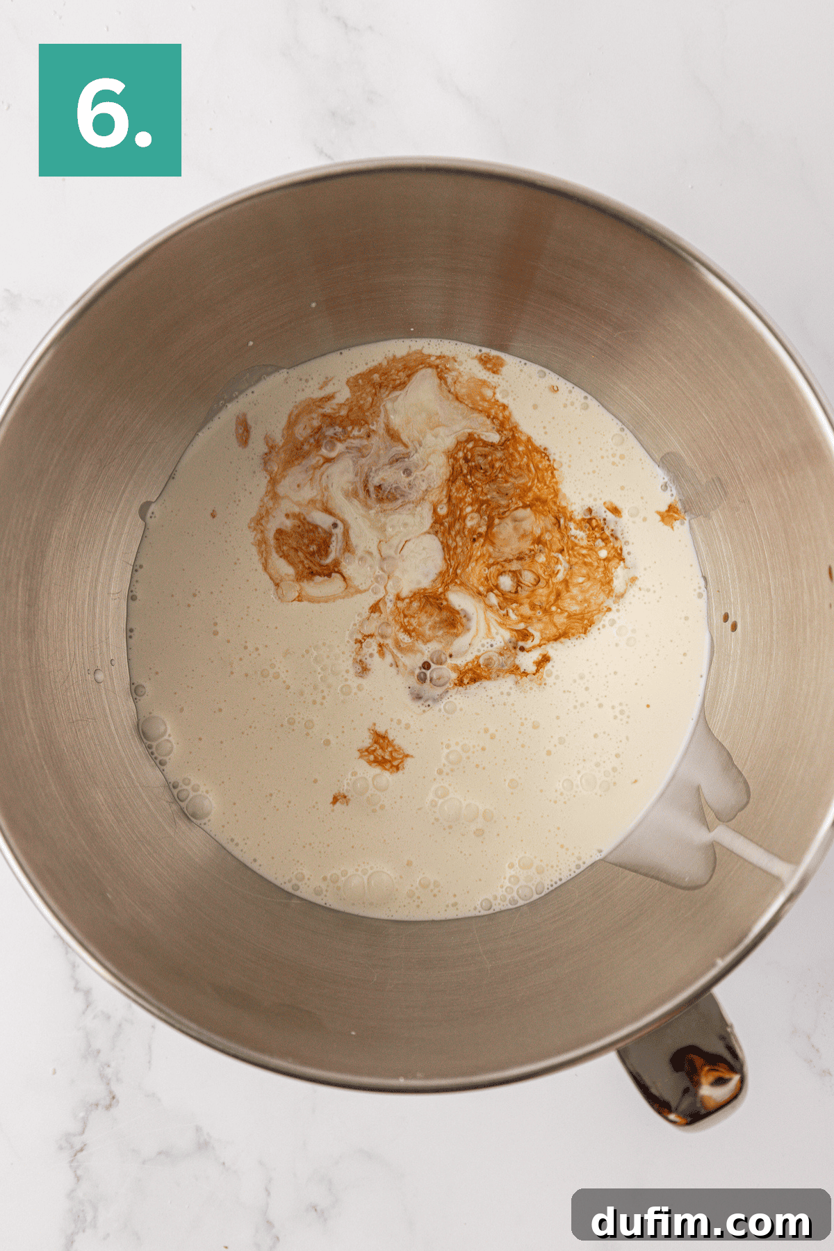 Whip Up a Philadelphia No Bake Cheesecake 11 A sleek metal mixing bowl, viewed from directly above, brimming with chilled heavy cream and fragrant vanilla extract, meticulously prepared for a Philadelphia no-bake cheesecake. The bowl rests on an elegant white marble surface, distinguished by a green box with the number '6' in the top-left corner of the image, marking a key stage.