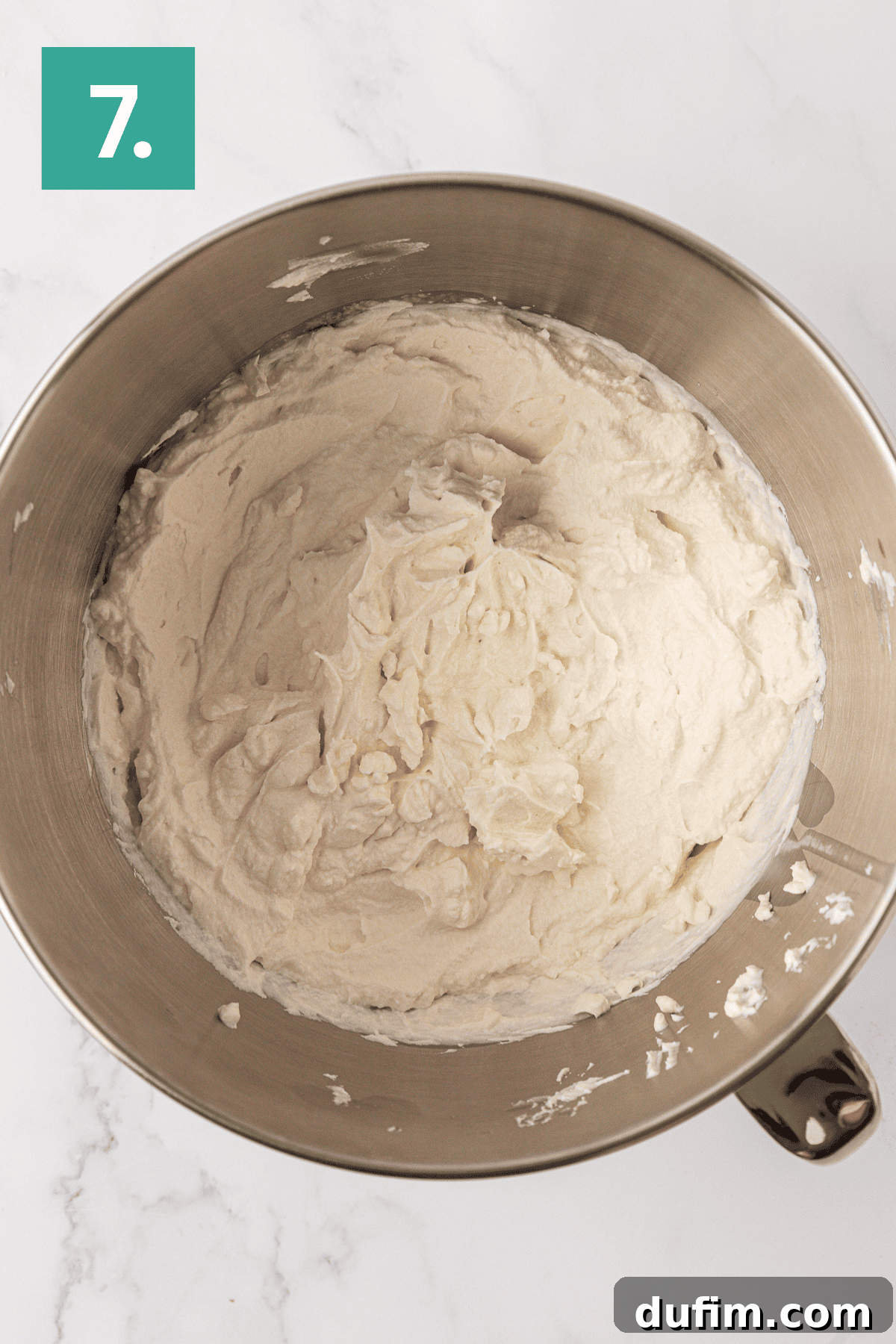Whip Up a Philadelphia No Bake Cheesecake 12 A gleaming metal mixing bowl holds a perfectly smooth and creamy Philadelphia no-bake cheesecake batter, artistically arranged on a pristine white marble surface. A bold green box in the upper left corner of the image proudly displays the number '7', signifying this crucial stage of the preparation.