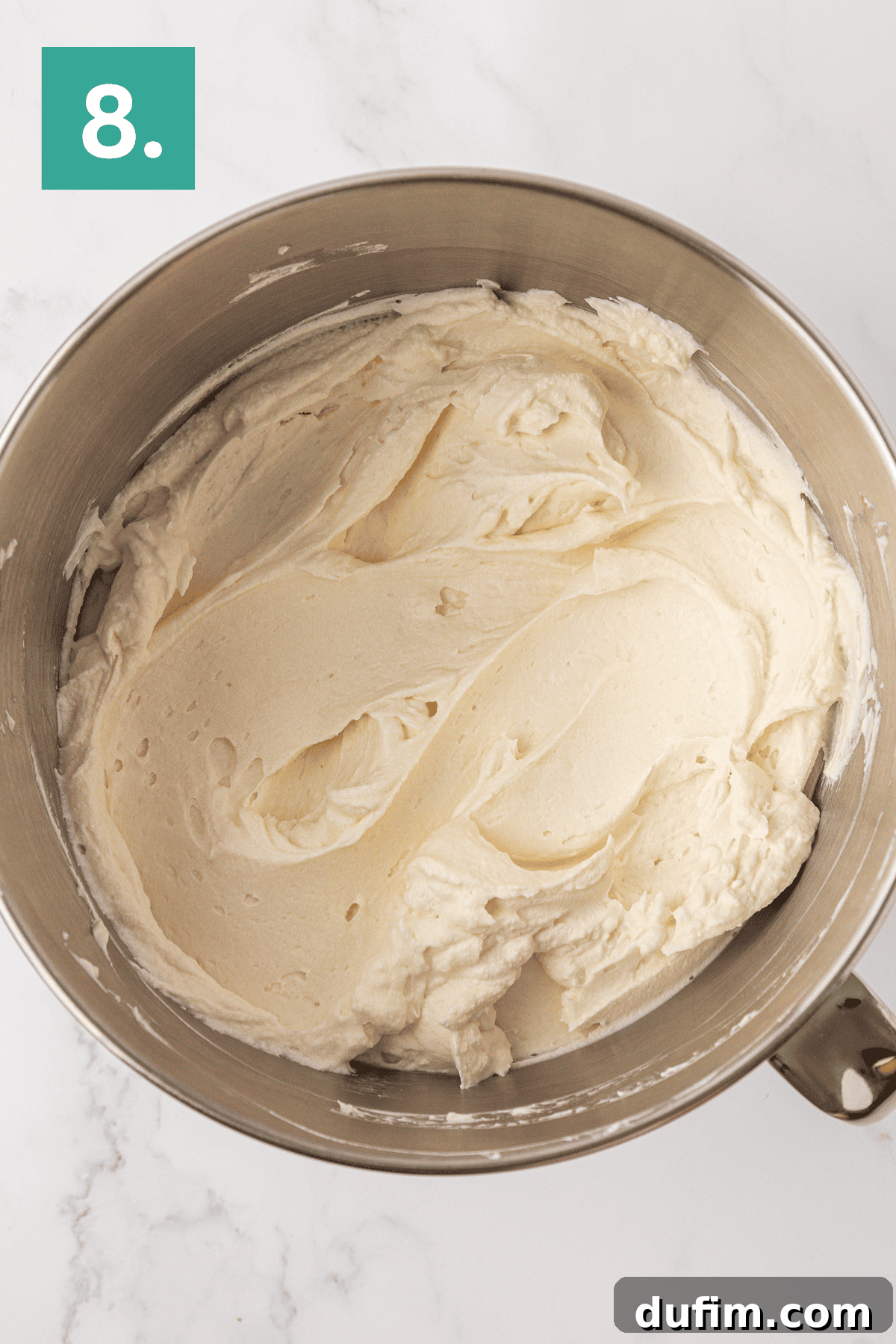 Whip Up a Philadelphia No Bake Cheesecake 13 A pristine mixing bowl filled with a luxuriously smooth, creamy white frosting, destined for a Philadelphia no-bake cheesecake, set against a backdrop of white marble. A prominent green box with the number '8' in white is positioned in the top-left corner, marking this pivotal stage in the dessert's creation.