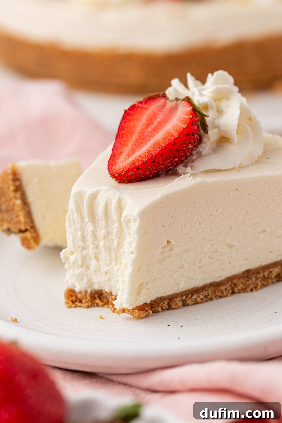 Whip Up a Philadelphia No Bake Cheesecake 15 A perfect, creamy slice of Philadelphia no-bake cheesecake with its distinct graham cracker crust, crowned with a succulent halved strawberry and a delicate swirl of whipped cream. The enticing dessert is artfully presented on a crisp white plate, set against a softly blurred background, inviting immediate indulgence and visual pleasure.
