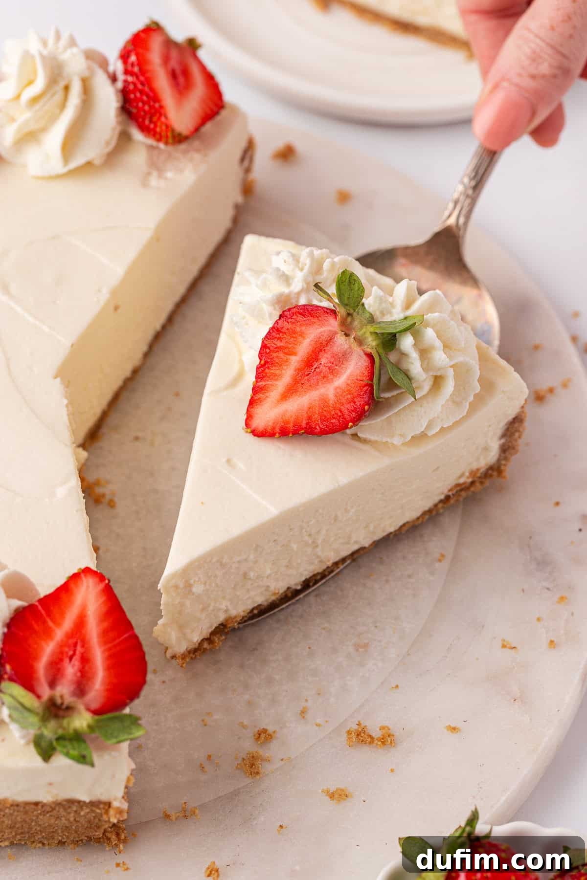 Whip Up a Philadelphia No Bake Cheesecake 16 A hand gracefully lifts a slice of exquisitely creamy Philadelphia no-bake cheesecake, crowned with a generous dollop of whipped cream and a vibrant halved strawberry, from a whole, inviting cheesecake. The scene is set on a pristine marble surface, with scattered crumbs and additional fresh strawberries nearby, creating an appetizing and lively tableau of dessert perfection.