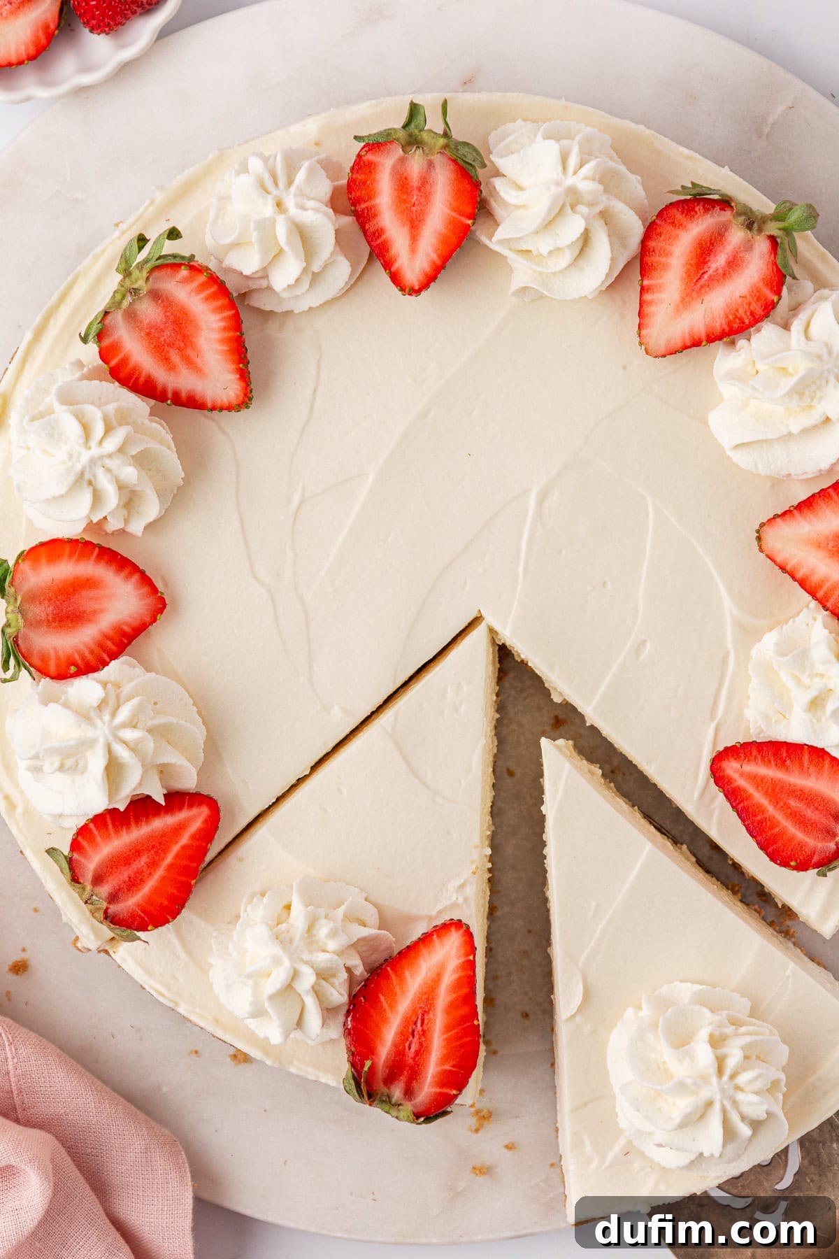 Whip Up a Philadelphia No Bake Cheesecake 3 A stunning round Philadelphia no-bake cheesecake, generously adorned with elegant swirls of homemade whipped cream and vibrant halved strawberries. One perfect slice is being carefully lifted out, revealing the creamy texture and inviting golden graham cracker crust. The cheesecake is presented on a clean white surface alongside a soft pink cloth, highlighting its fresh appeal.
