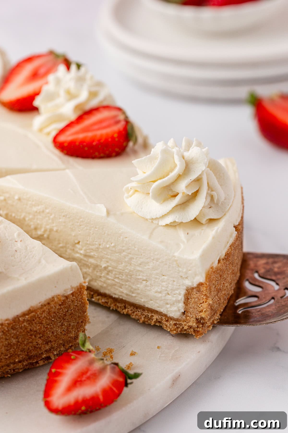 Whip Up a Philadelphia No Bake Cheesecake 5 A delectable slice of creamy Philadelphia no-bake cheesecake, featuring a perfectly crumbly golden graham cracker crust. The slice is beautifully adorned with generous swirls of whipped cream and a fresh halved strawberry, presented elegantly on a pristine white surface, ready to be enjoyed as a delightful dessert.