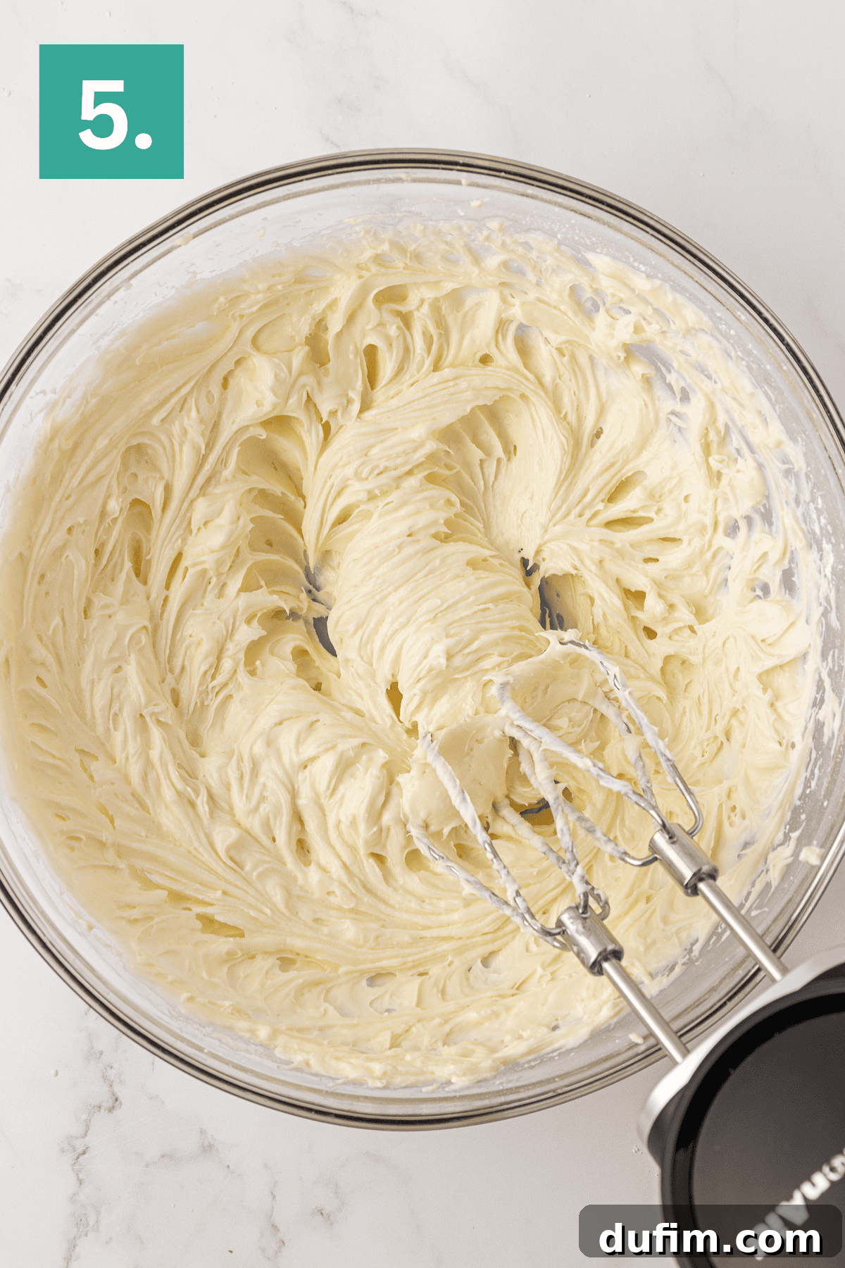 Whip Up a Philadelphia No Bake Cheesecake 10 A creamy, smooth whipped cream cheese mixture, destined for a Philadelphia no-bake cheesecake, is actively being blended in a glass bowl with an electric hand mixer. A distinct green box in the upper left corner prominently displays the number '5.', indicating this crucial step in the preparation process.