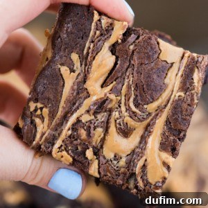 A hand with light blue nail polish holds a thick, rich chocolate brownie featuring luscious peanut butter swirl brownies and visible swirls of creamy peanut butter.