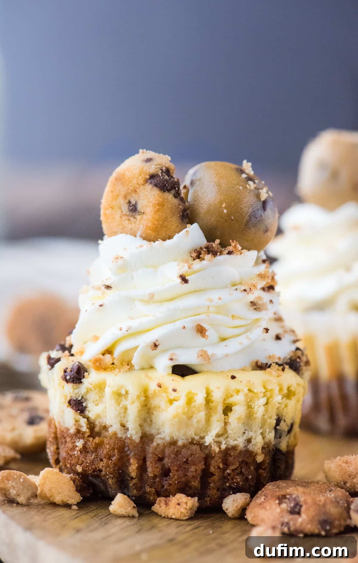 A beautifully presented mini chocolate chip cookie dough cheesecake with a golden-brown cookie crust, topped with generous swirls of whipped cream, scattered mini chocolate chips, and a small cookie dough ball, all resting on a rustic wooden tray.