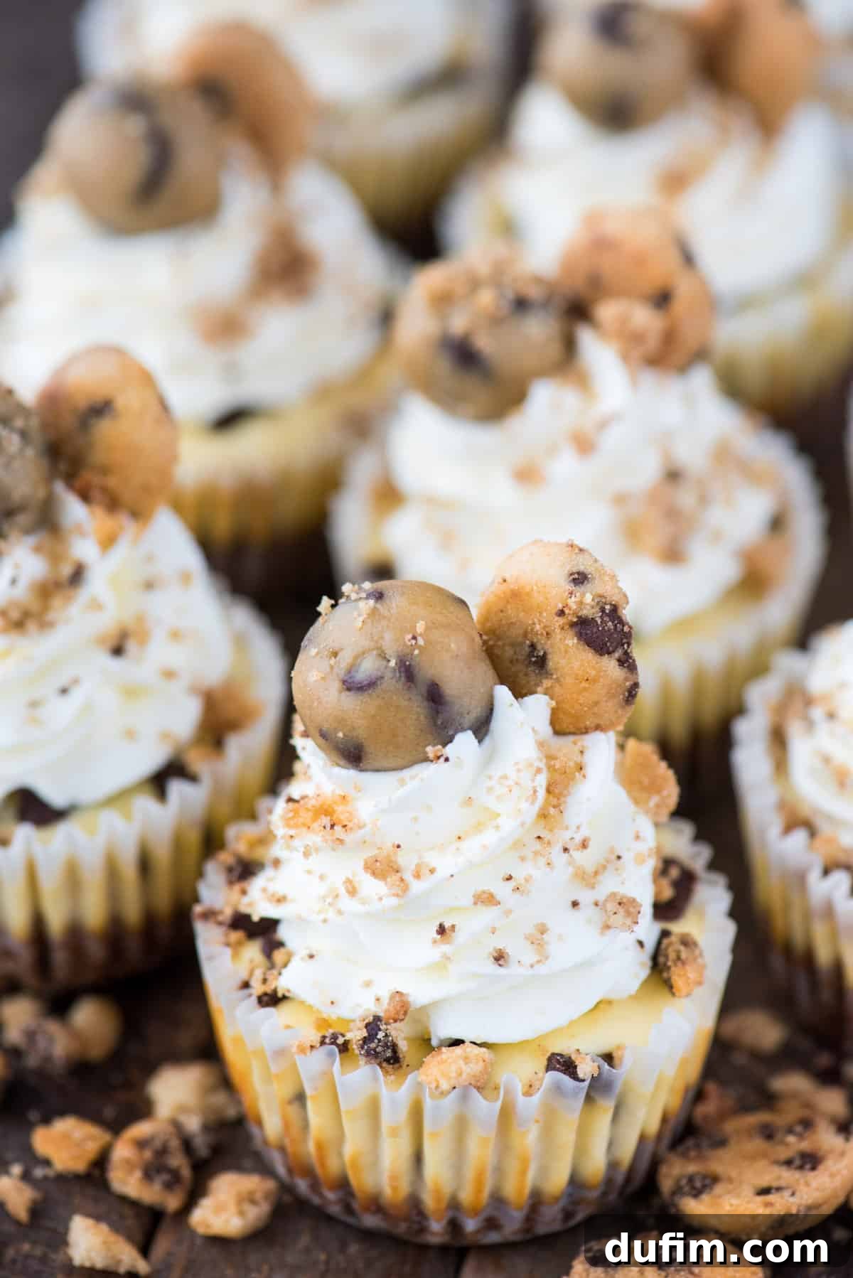 An array of mini chocolate chip cookie dough cheesecakes, each featuring a golden cookie crust and a creamy cheesecake topping studded with mini chocolate chips, presented on a dark, elegant background.