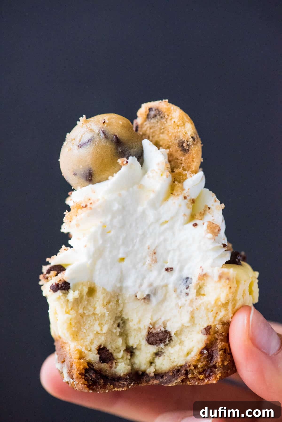 A close-up shot of a hand holding a mini chocolate chip cookie dough cheesecake with a bite removed, revealing the creamy filling and cookie crust, set against a blurred background.
