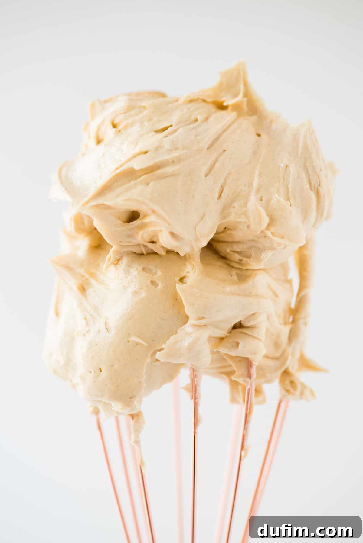 Decadent Peanut Butter Fluff 5 The perfect consistency of creamy peanut butter frosting clinging to a rose gold whisk, against a clean white background, highlighting its smooth and pipeable texture.