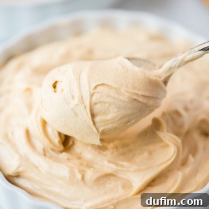 A spoon scoops rich, creamy peanut butter frosting from a white, fluted dish brimming with light brown peanut butter frosting.