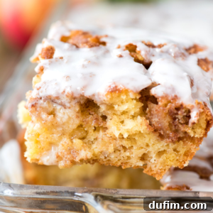 A close-up of a piece of cinnamon roll cake with a moist, fluffy texture and swirled cinnamon, reminiscent of apple coffee cake, topped with a generous layer of white icing.