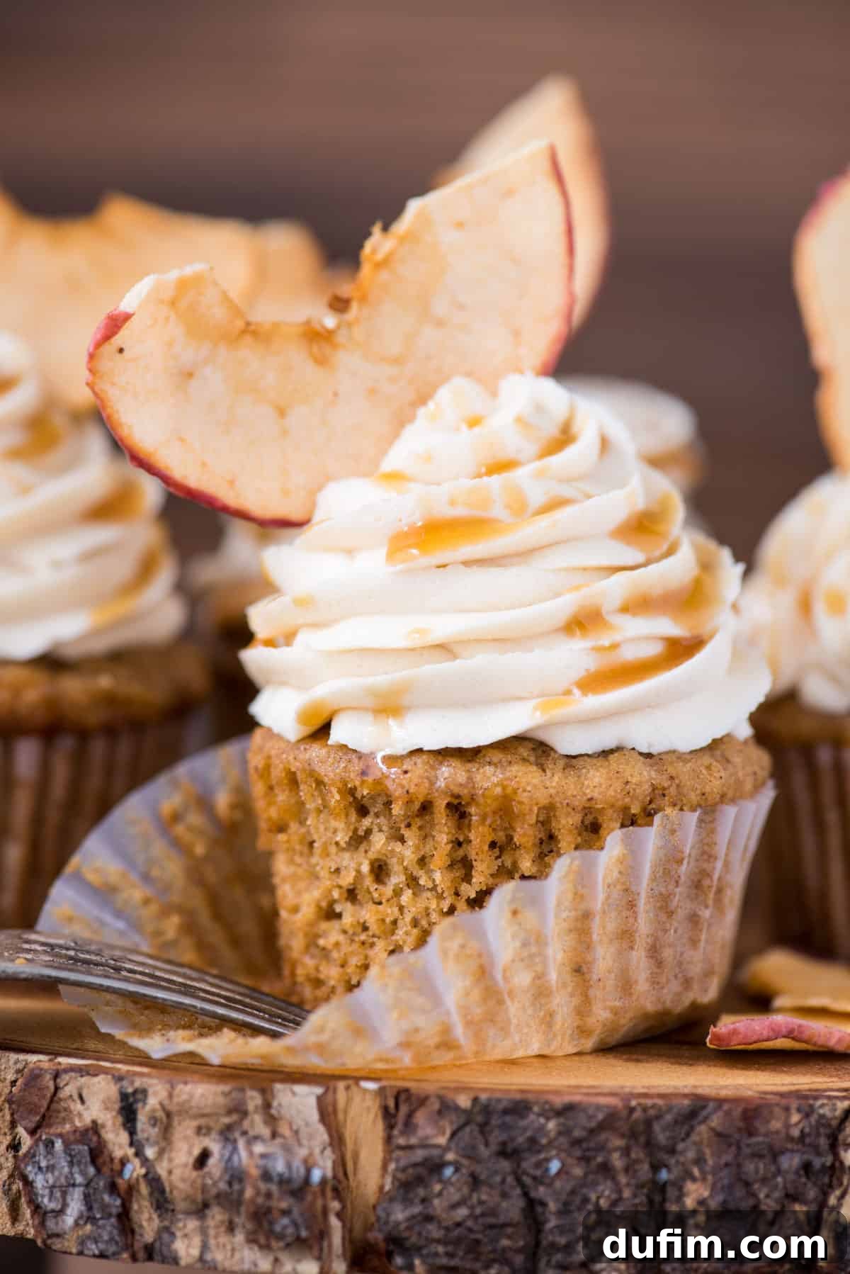 Spiced Apple Harvest Cupcakes 3 Close-up of a single apple spice cupcake with fluffy salted caramel frosting and a delicate apple chip garnish, resting on a wooden cake stand with a blurred rustic background.