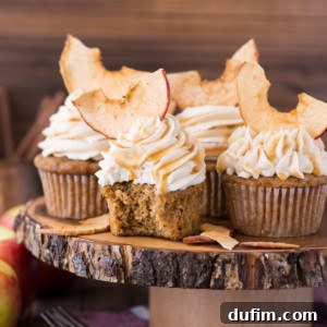 Three apple spice cupcakes with swirled salted caramel frosting and dried apple slices on top are displayed on a rustic wooden stand. An inviting bite is taken from one cupcake, revealing its moist interior. Apples and forks are in the blurred background, enhancing the autumn theme.