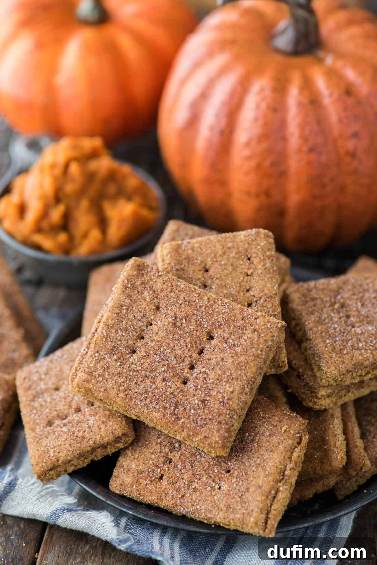 Pumpkin Spice Graham Snaps 2 A generous pile of homemade pumpkin graham crackers in a rustic metal bowl, with autumnal pumpkins and foliage blurred in the background, creating a warm and inviting scene.