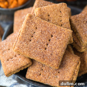 A close-up of a plate piled with rectangular, cinnamon-sugar-dusted pumpkin graham crackers, each with small holes pricked in the center. Some crackers are stacked, showing their crisp, golden-brown texture.