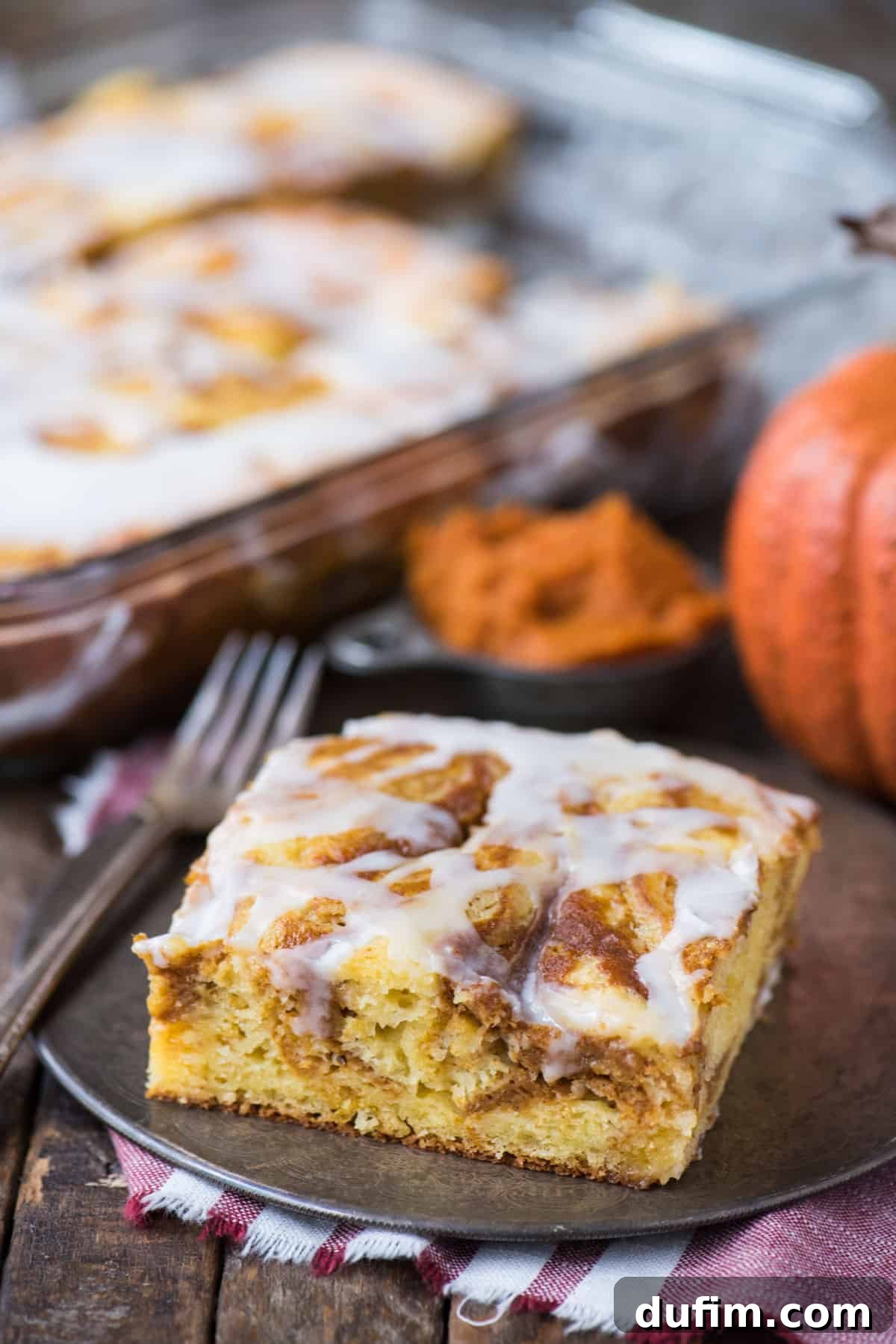 A pristine slice of easy pumpkin swirl cake, adorned with a delicate white icing, elegantly presented on a metal plate. The internal swirls of pumpkin are clearly visible against the yellow cake base.