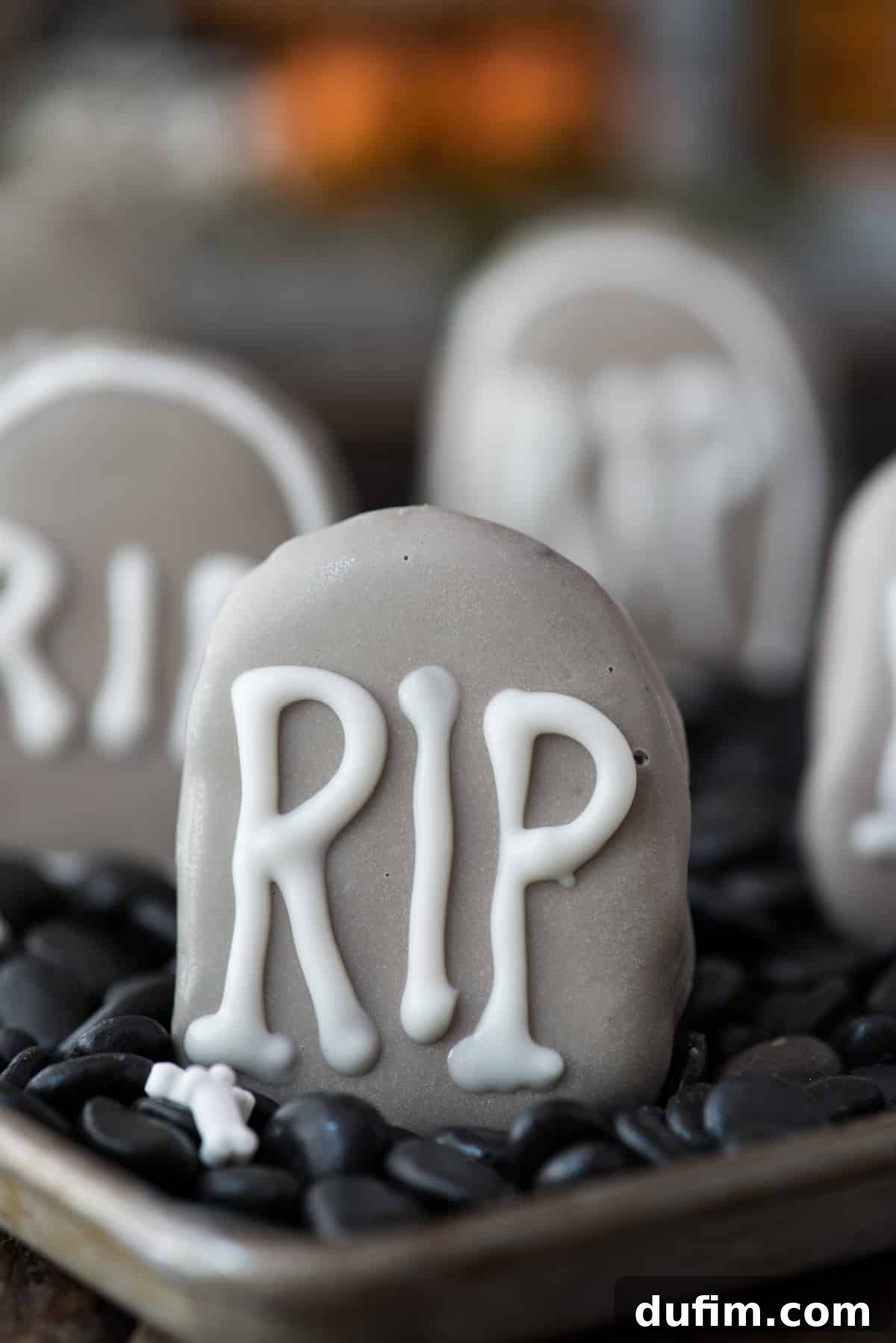 Two spooky tombstone-shaped Oreo balls nestled in a shallow pan, surrounded by black decorative rocks, creating a Halloween graveyard scene. One tombstone has 'RIP' written on it in white icing.