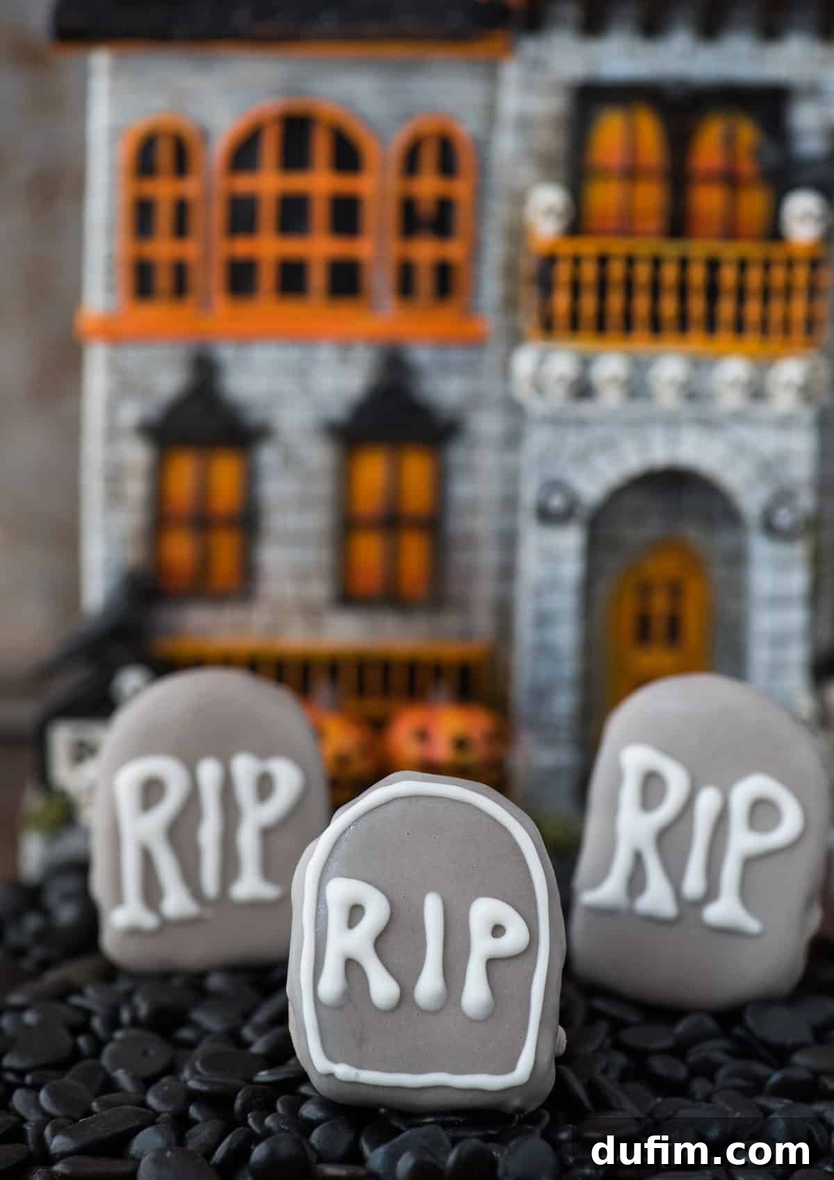 A collection of tombstone-shaped Oreo balls in a shallow pan, surrounded by dark stones, with a haunted house decoration subtly visible in the soft-focus background, enhancing the spooky Halloween atmosphere.