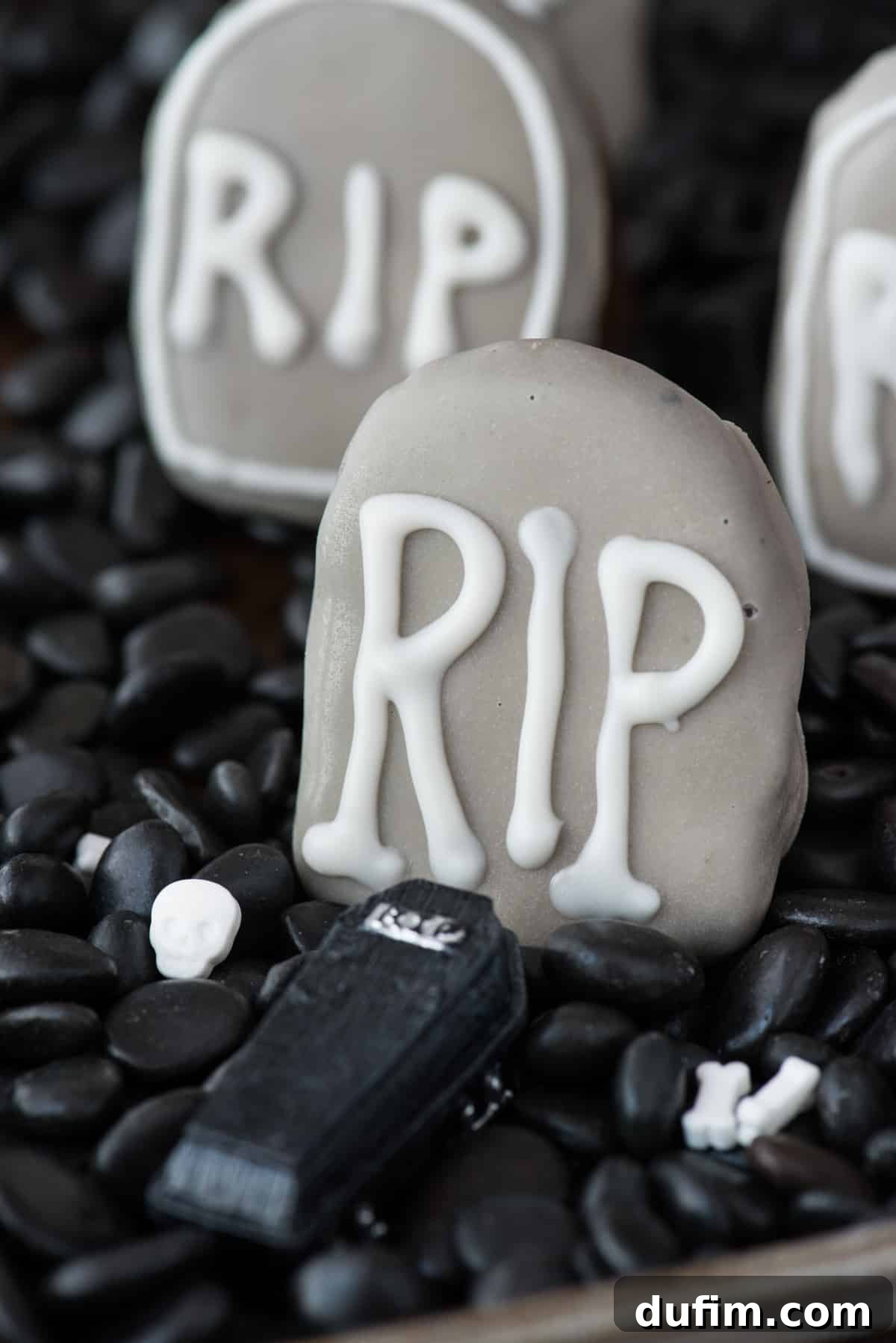 A single, perfectly dipped tombstone-shaped Oreo ball with 'RIP' in white icing, resting on a bed of black rocks. The lighting highlights its gray coating and neat details, emphasizing its spooky elegance.