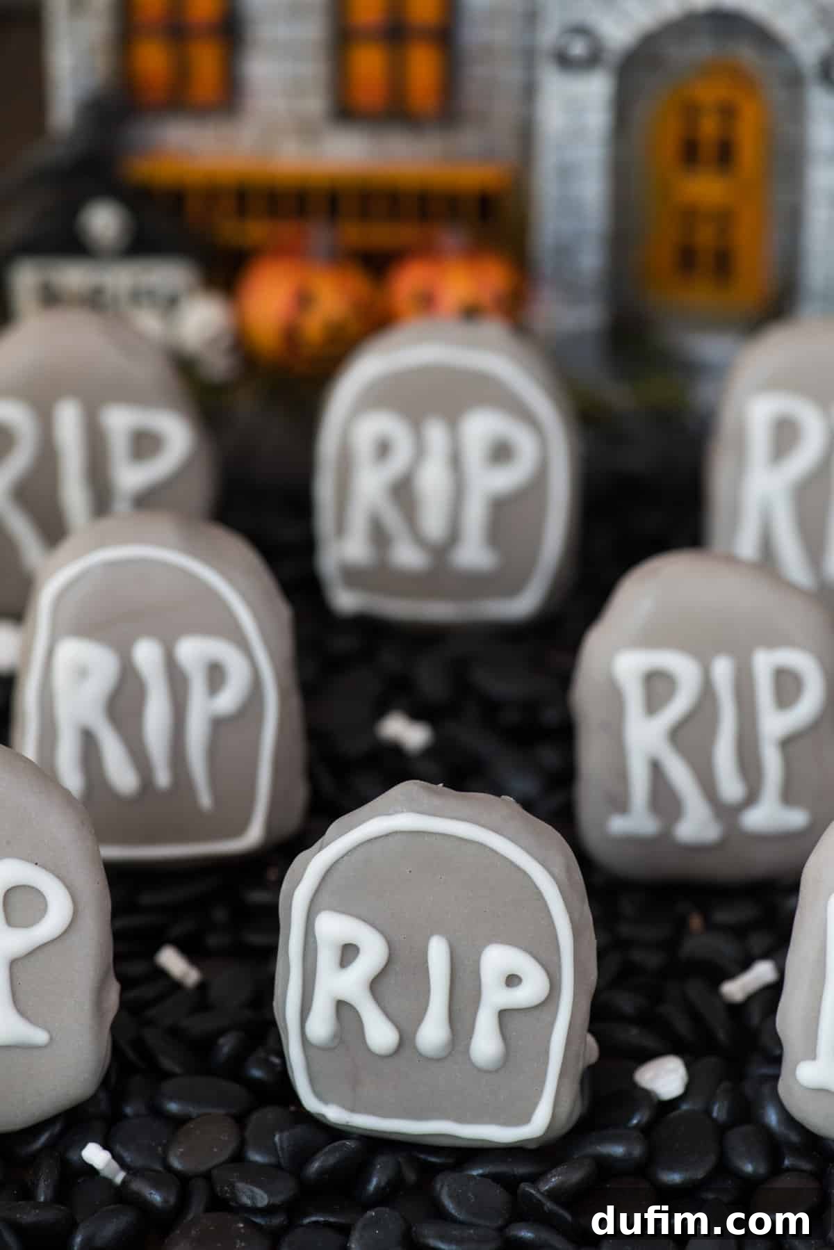 A creative graveyard display of tombstone-shaped Oreo balls, arranged in a shallow pan with black rocks and a haunted house decoration in the background. The spooky setup is perfect for a Halloween party, creating a fun yet eerie atmosphere.