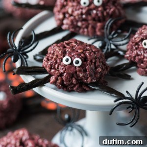 Halloween rice krispie treats shaped like spiders, with chocolate, candy eyes, black licorice legs, and plastic spider decorations, are displayed on a white cake stand.