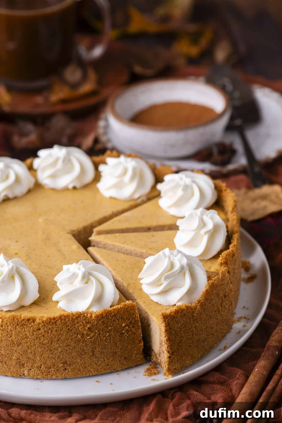 A pumpkin cheesecake with a graham cracker crust, topped with dollops of whipped cream, sits on a plate with a slice removed. Nearby are a bowl of cinnamon, a cup of coffee, and autumn leaves.