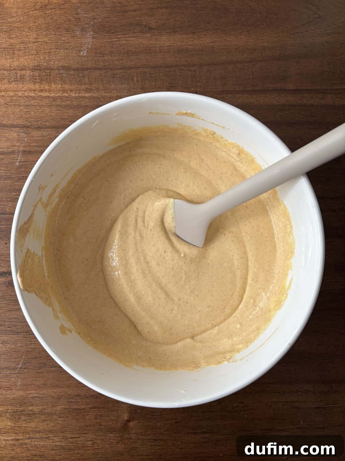 A white mixing bowl filled with light brown batter and a white spatula resting inside, placed on a wooden surface.
