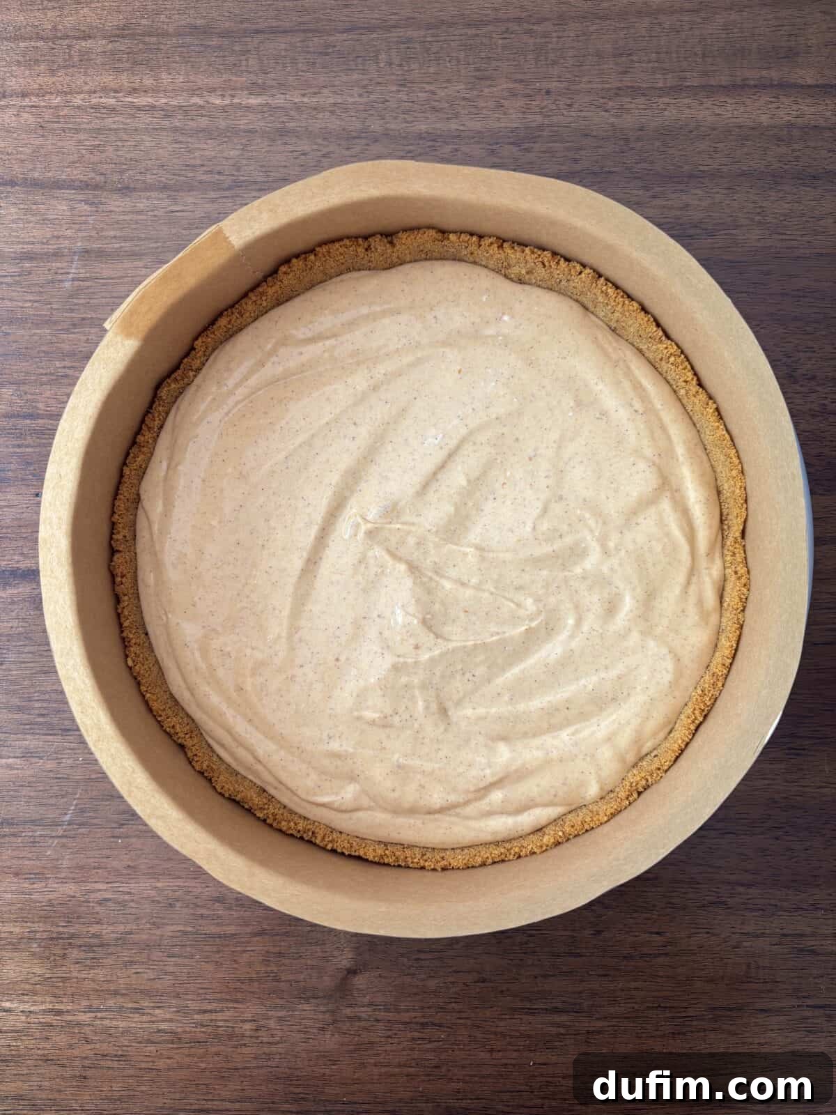 A round pie with a graham cracker crust and a smooth, creamy tan filling sits in a parchment-lined baking tin on a wooden surface.