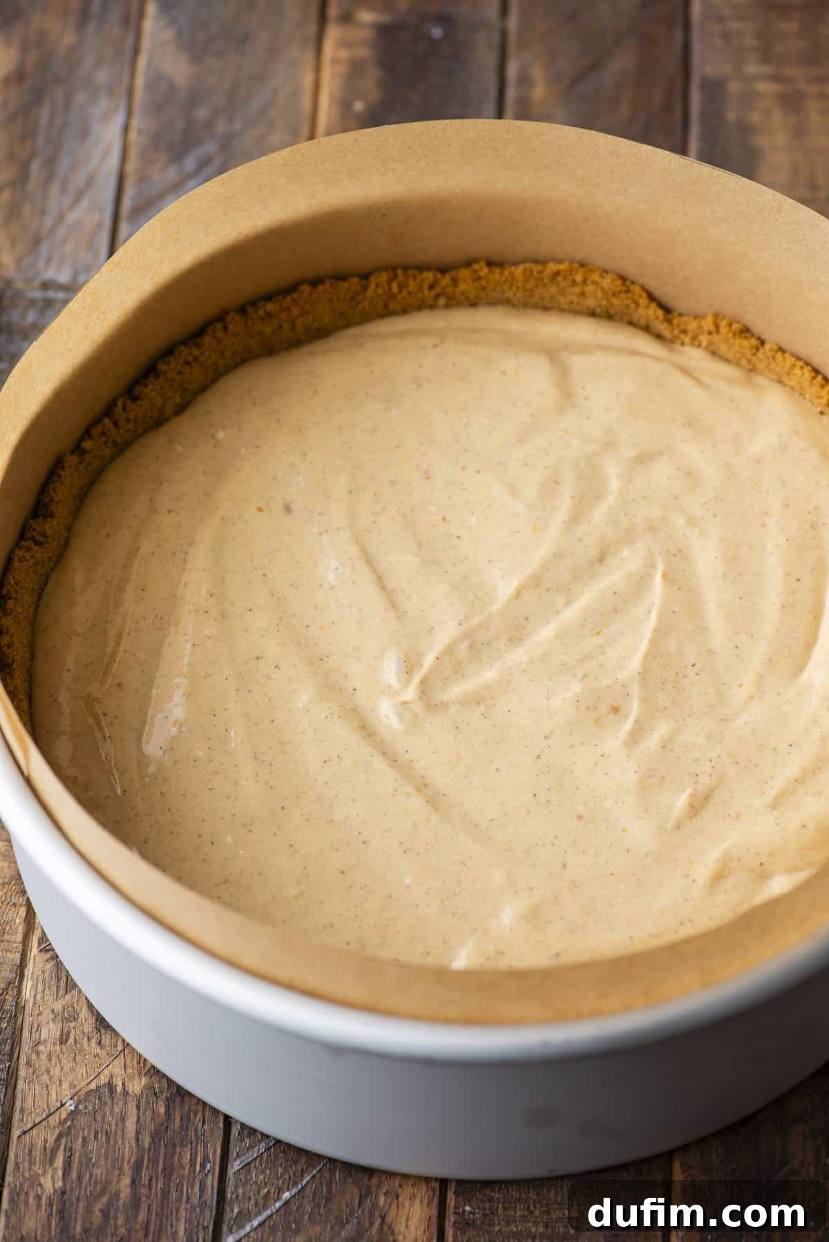 A round baking pan lined with parchment paper, filled with a smooth, creamy batter and a graham cracker crust along the edge, sits on a wooden surface.
