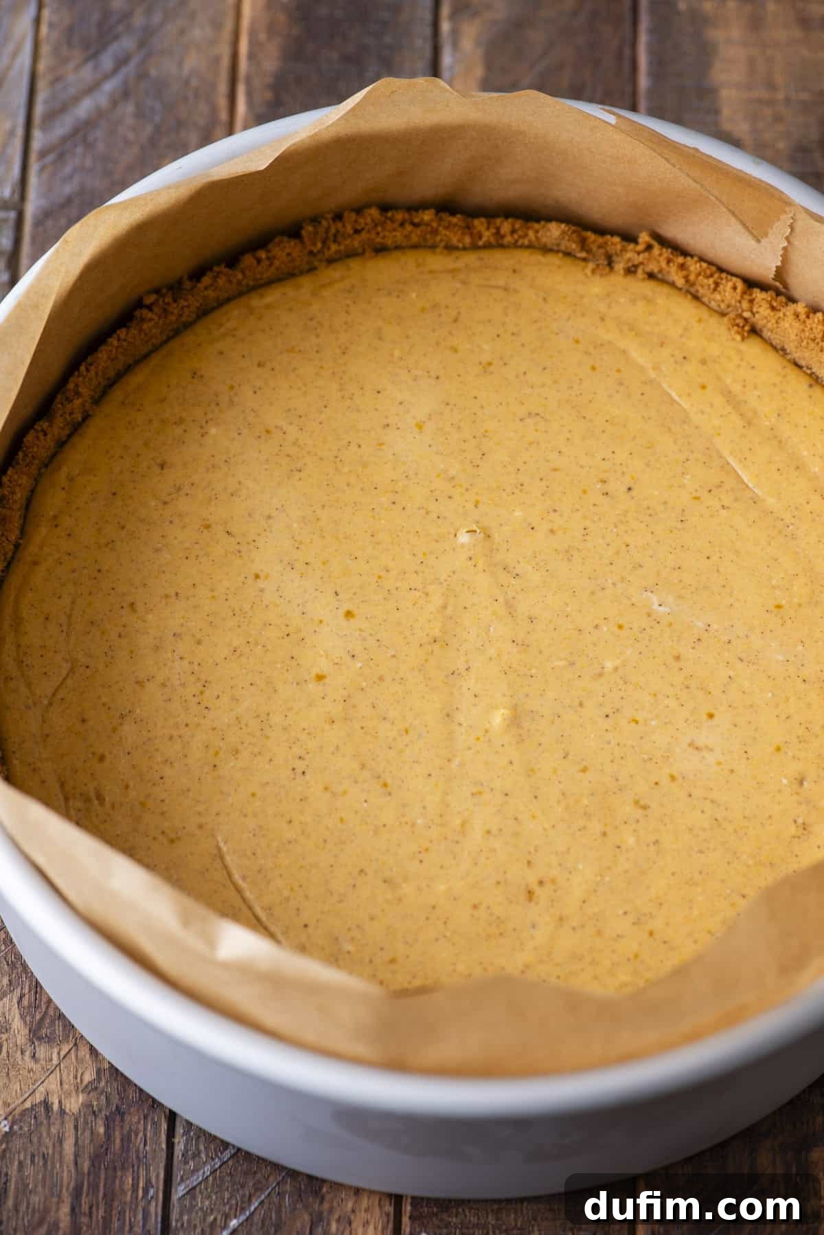 A round cheesecake with a graham cracker crust, lined with parchment paper, sits in a baking pan on a wooden surface. The filling is smooth and lightly golden, suggesting a pumpkin or spiced flavor.