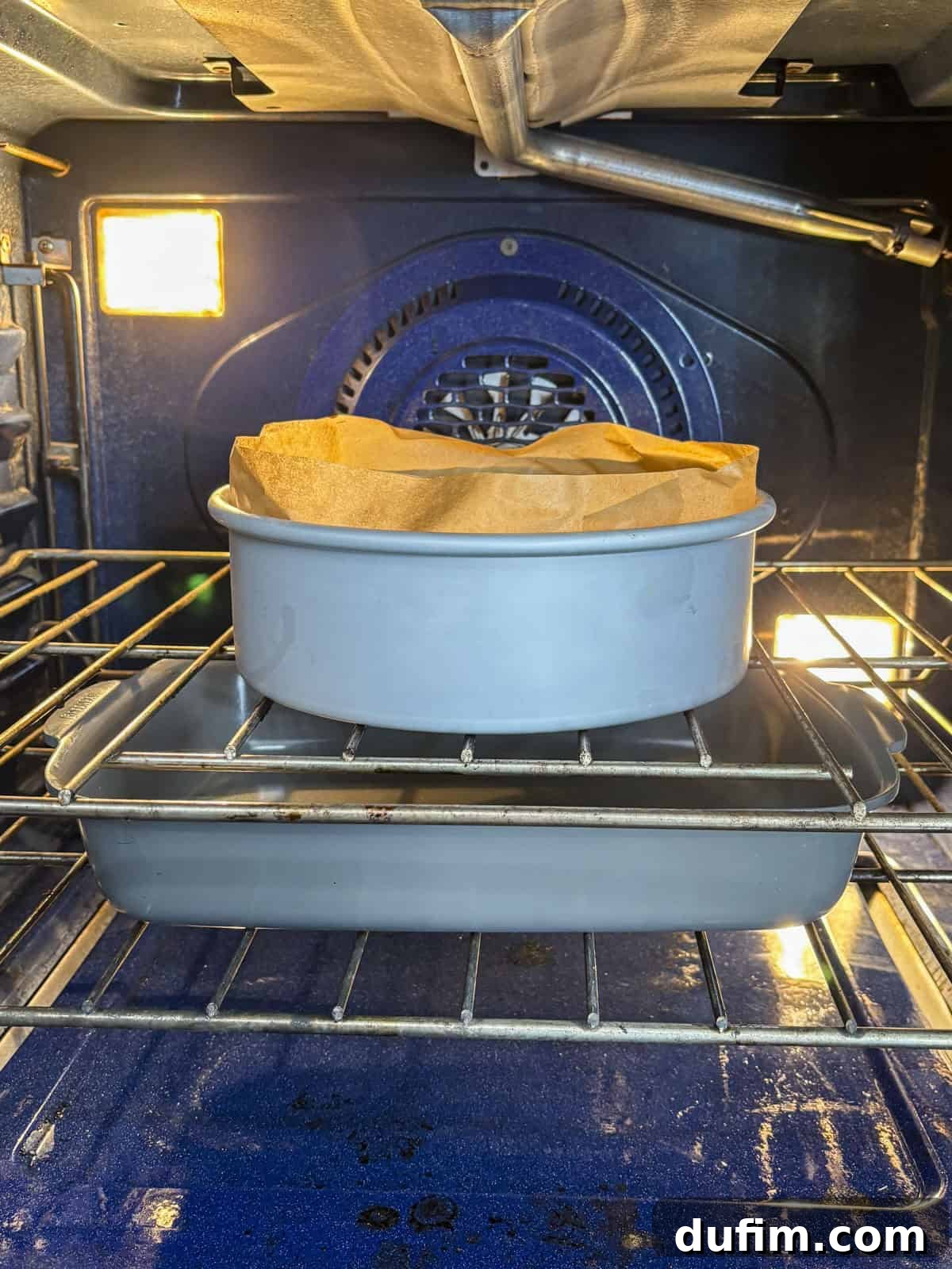 A round cake pan lined with parchment paper sits on a rectangular baking dish in an oven, with the oven light on and racks visible.
