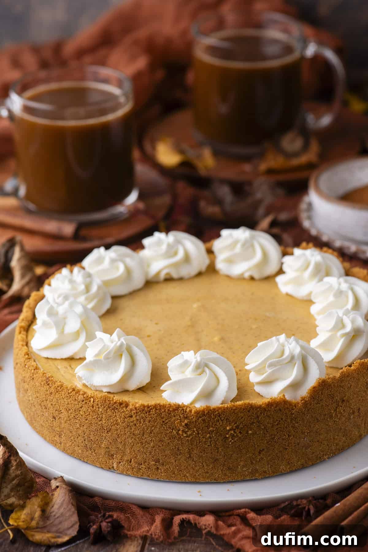 A pumpkin cheesecake with a graham cracker crust, topped with swirls of whipped cream, sits on a white plate. Two cups of coffee and autumn leaves are in the background.