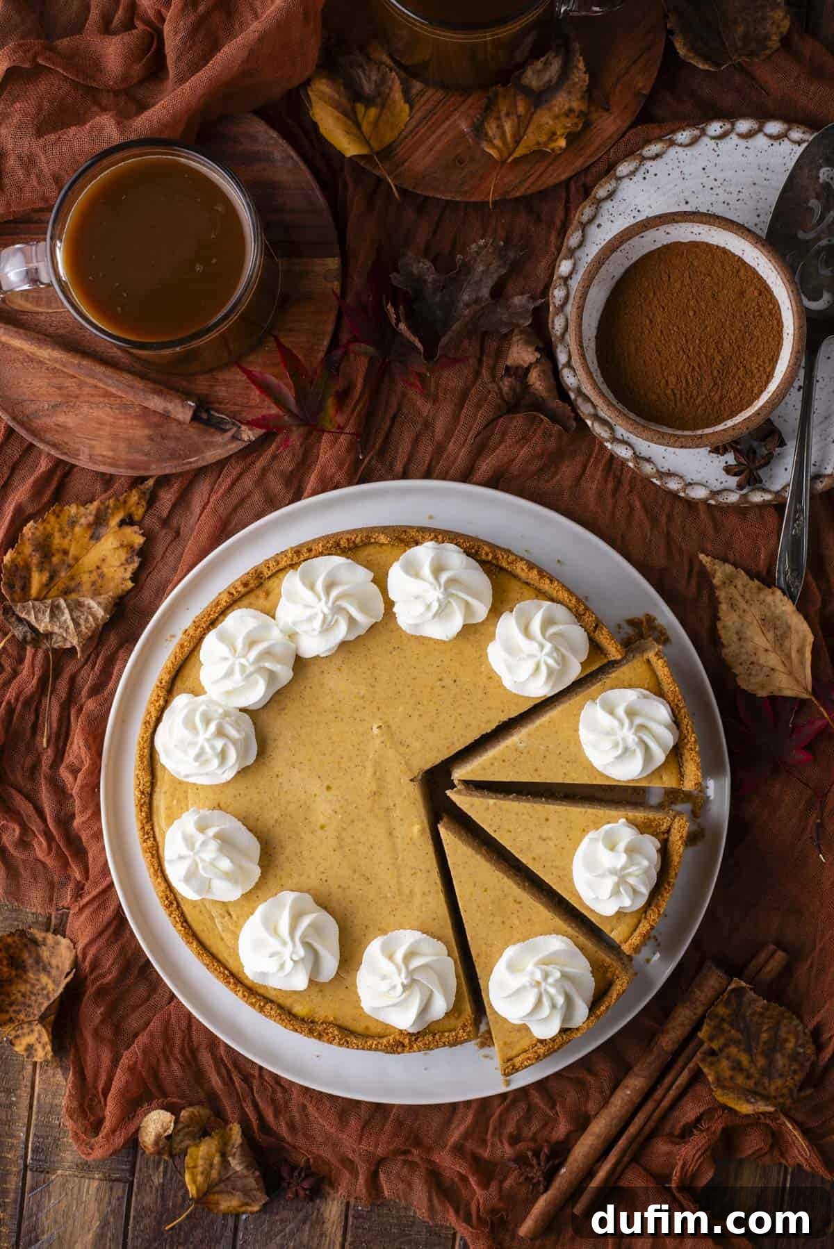 A whole pumpkin pie with whipped cream dollops on top, two slices cut, surrounded by a cup of coffee, bowl of cinnamon, fall leaves, and a rust-colored cloth on a wooden table.