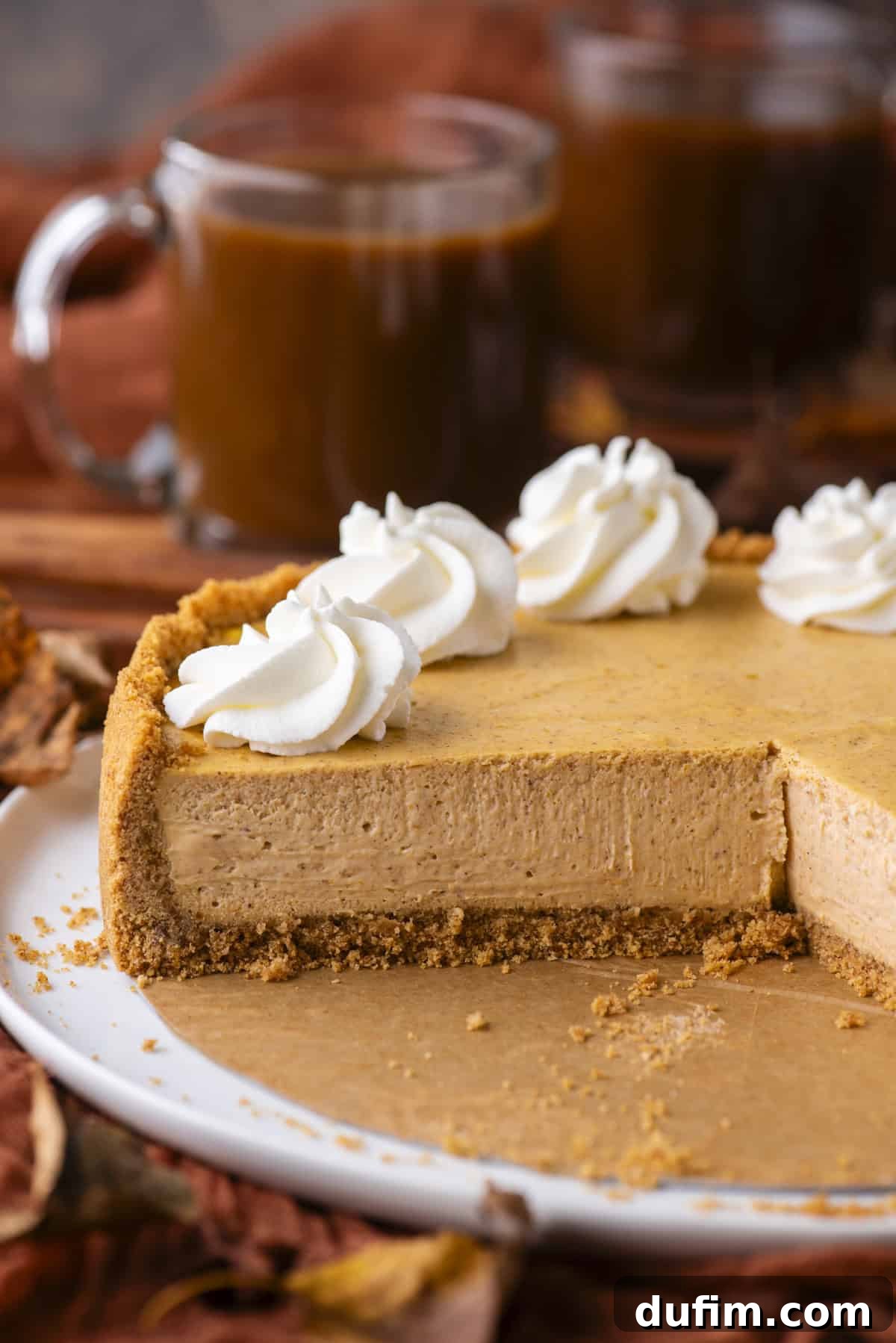 A pumpkin cheesecake with a graham cracker crust sits on a white plate. A slice has been removed, revealing a creamy filling. The cheesecake is topped with whipped cream swirls, and coffee mugs are in the background.