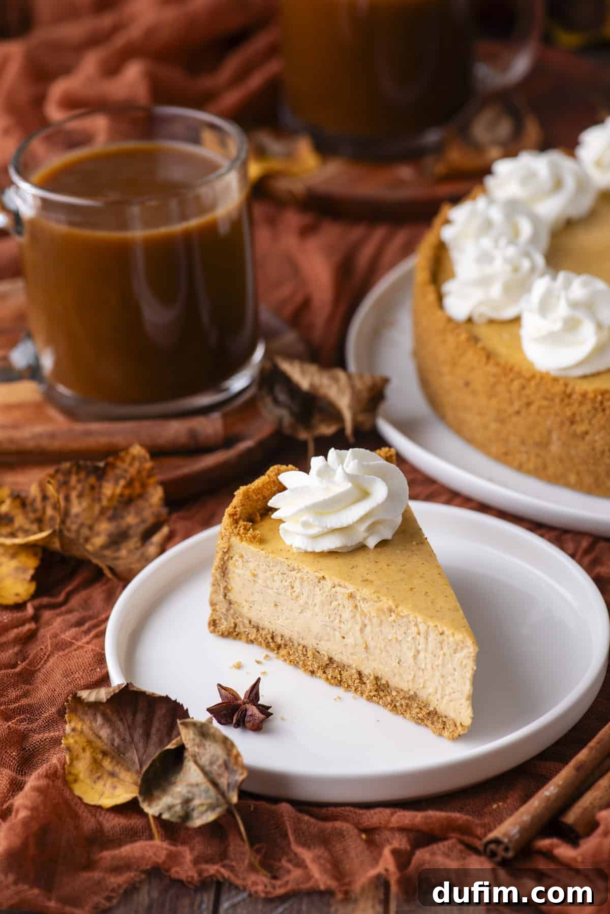 A slice of pumpkin cheesecake topped with whipped cream on a white plate, surrounded by autumn leaves, spices, and a glass mug of coffee. The whole cheesecake and another mug are visible in the background.