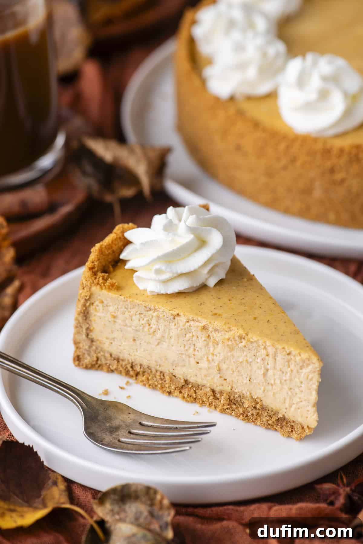 A slice of pumpkin cheesecake with a graham cracker crust and a swirl of whipped cream on top, served on a white plate with a fork. The whole cheesecake and autumn leaves are in the background.