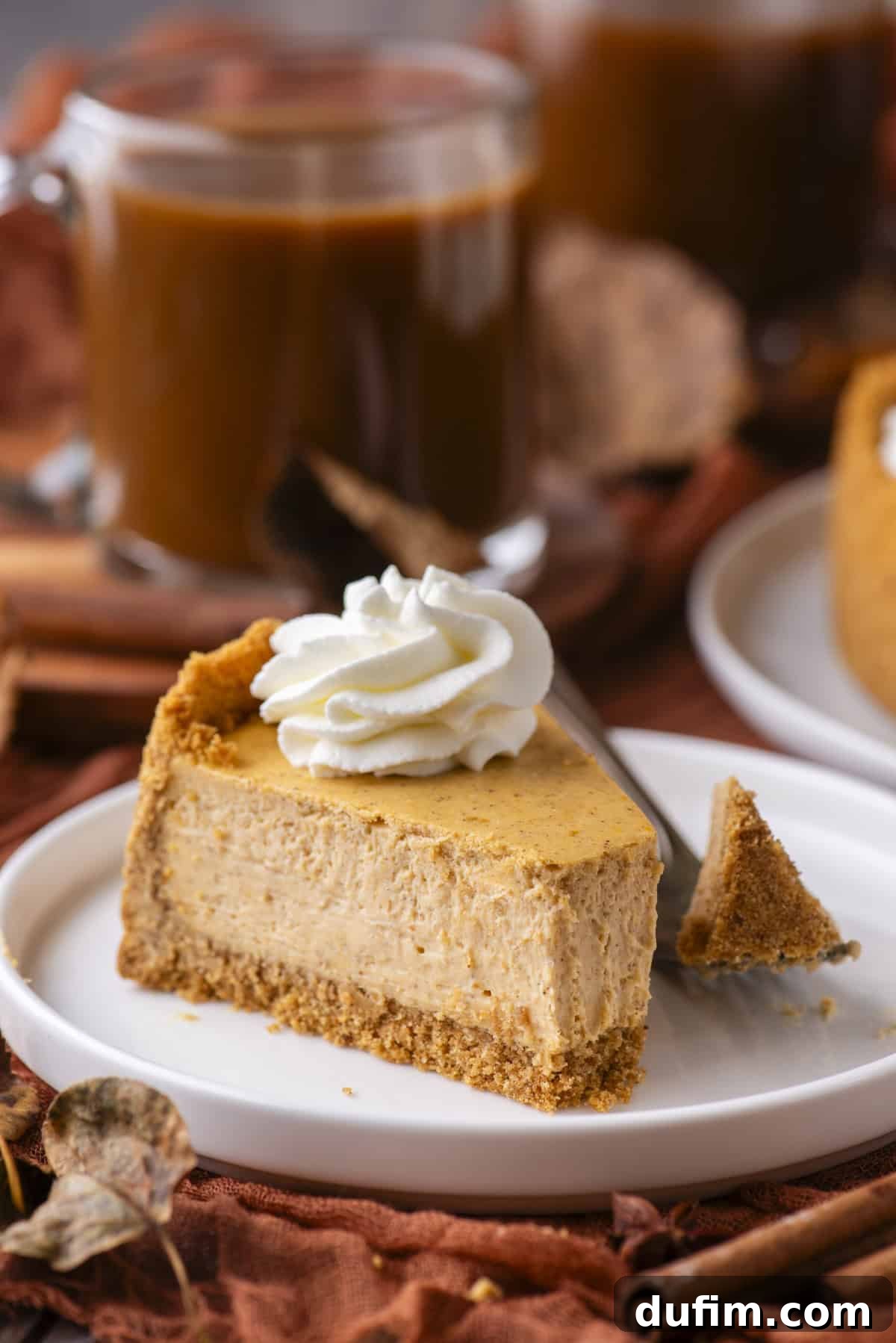 A slice of pumpkin cheesecake with a graham cracker crust and whipped cream topping sits on a white plate. A fork holds a bite of cheesecake nearby. In the background, there’s a glass mug of coffee and autumn decor.