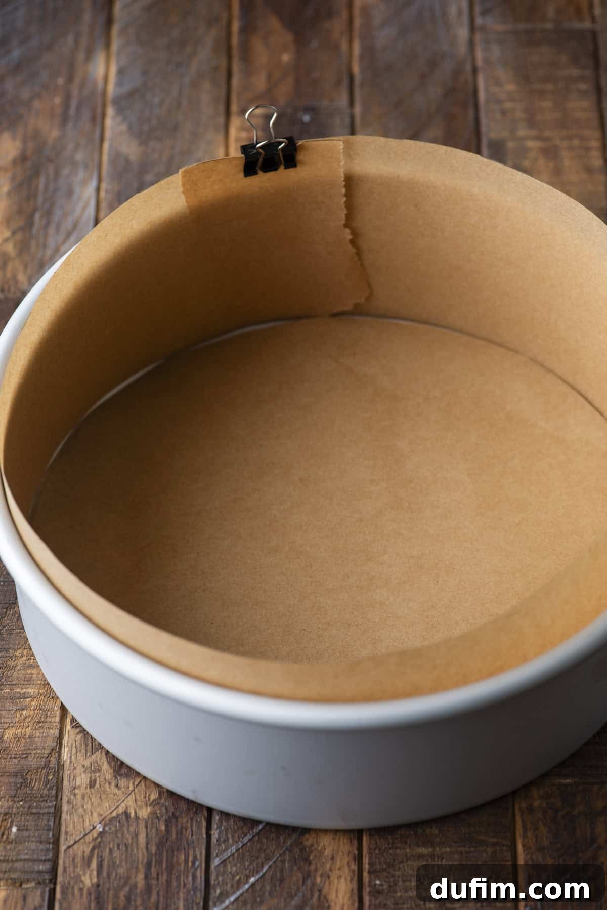 A round cake pan lined with brown parchment paper along the bottom and sides, held in place with a black binder clip, sits on a rustic wooden surface.