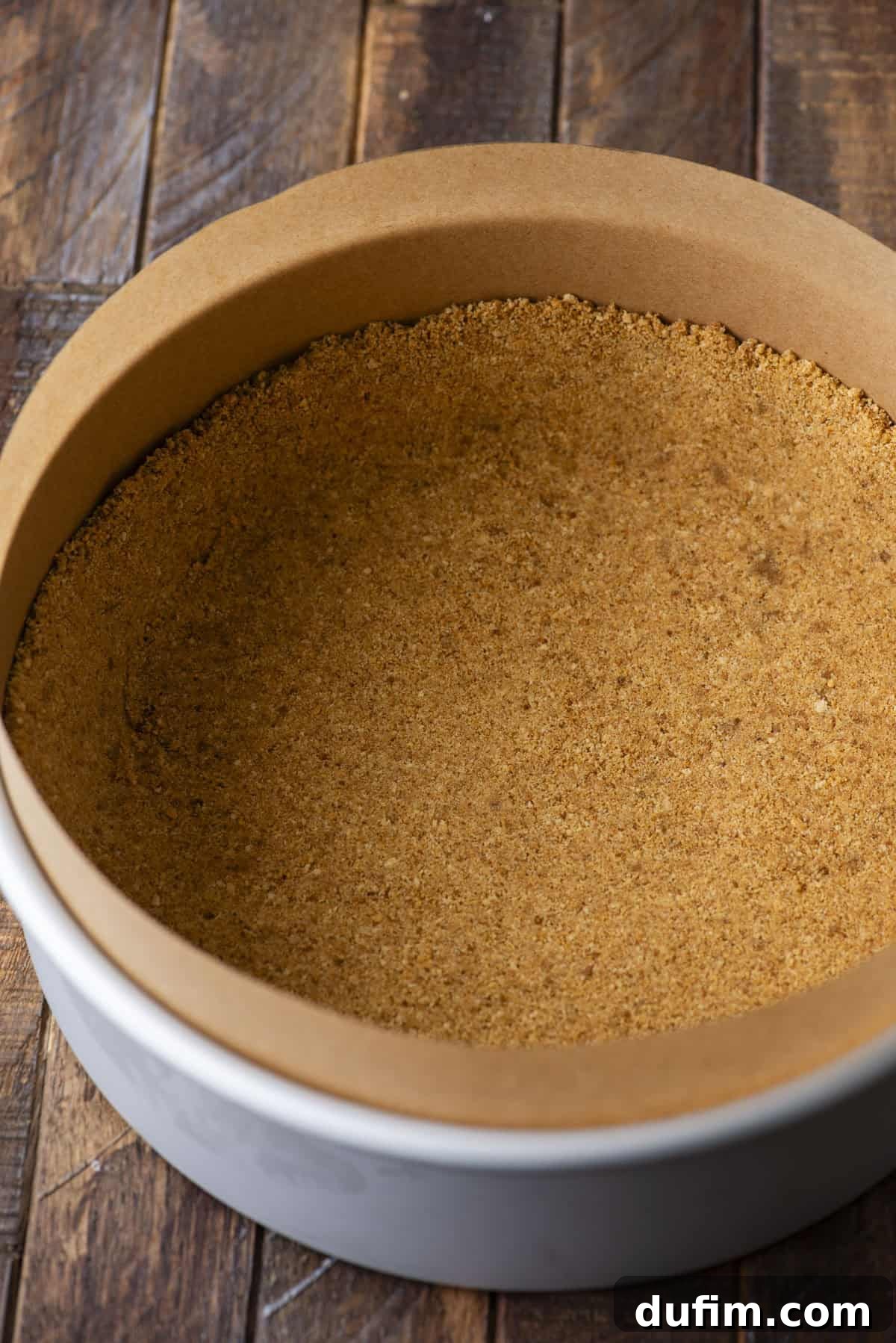 A round baking pan lined with parchment paper and filled with a pressed graham cracker crust, set on a rustic wooden surface.
