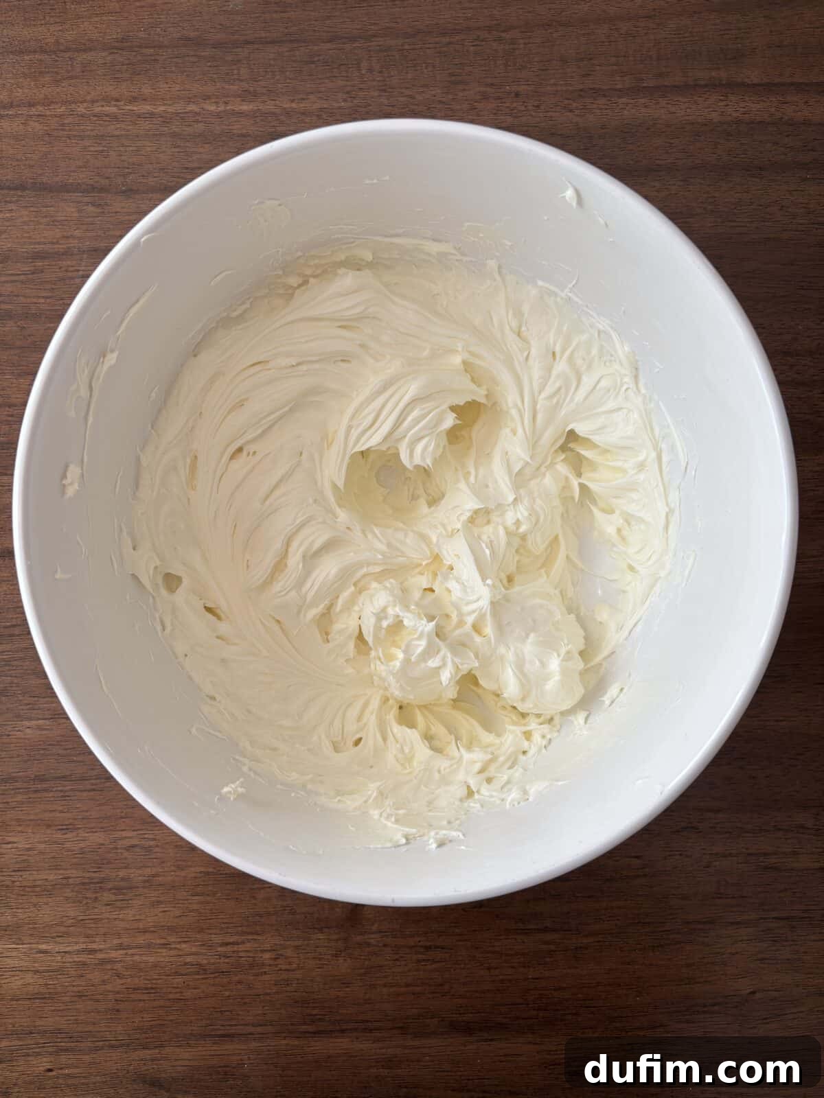 A large white mixing bowl filled with creamy, whipped frosting or batter sits on a dark wooden surface. The frosting has a smooth and fluffy texture with visible swirls from mixing.