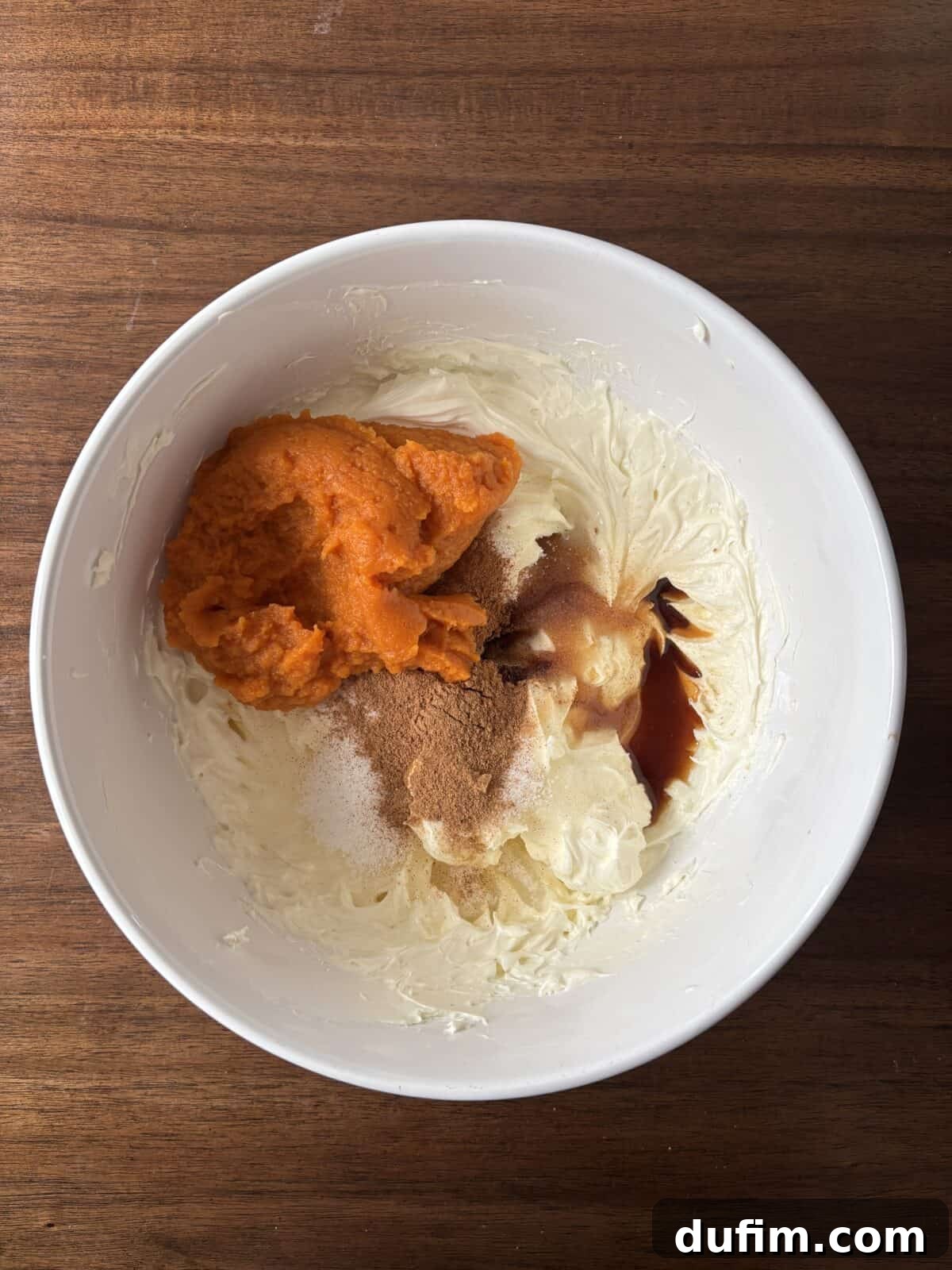 A white bowl on a wooden surface contains cream cheese, pumpkin puree, brown sugar, vanilla extract, and spices, ready to be mixed together.