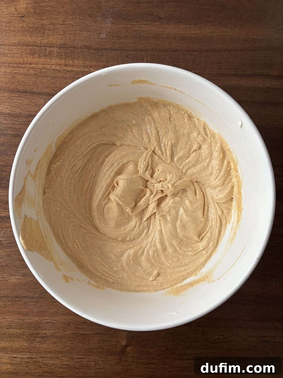 A white mixing bowl containing a light brown, creamy batter sits on a wooden surface. The batter has visible swirls from mixing.