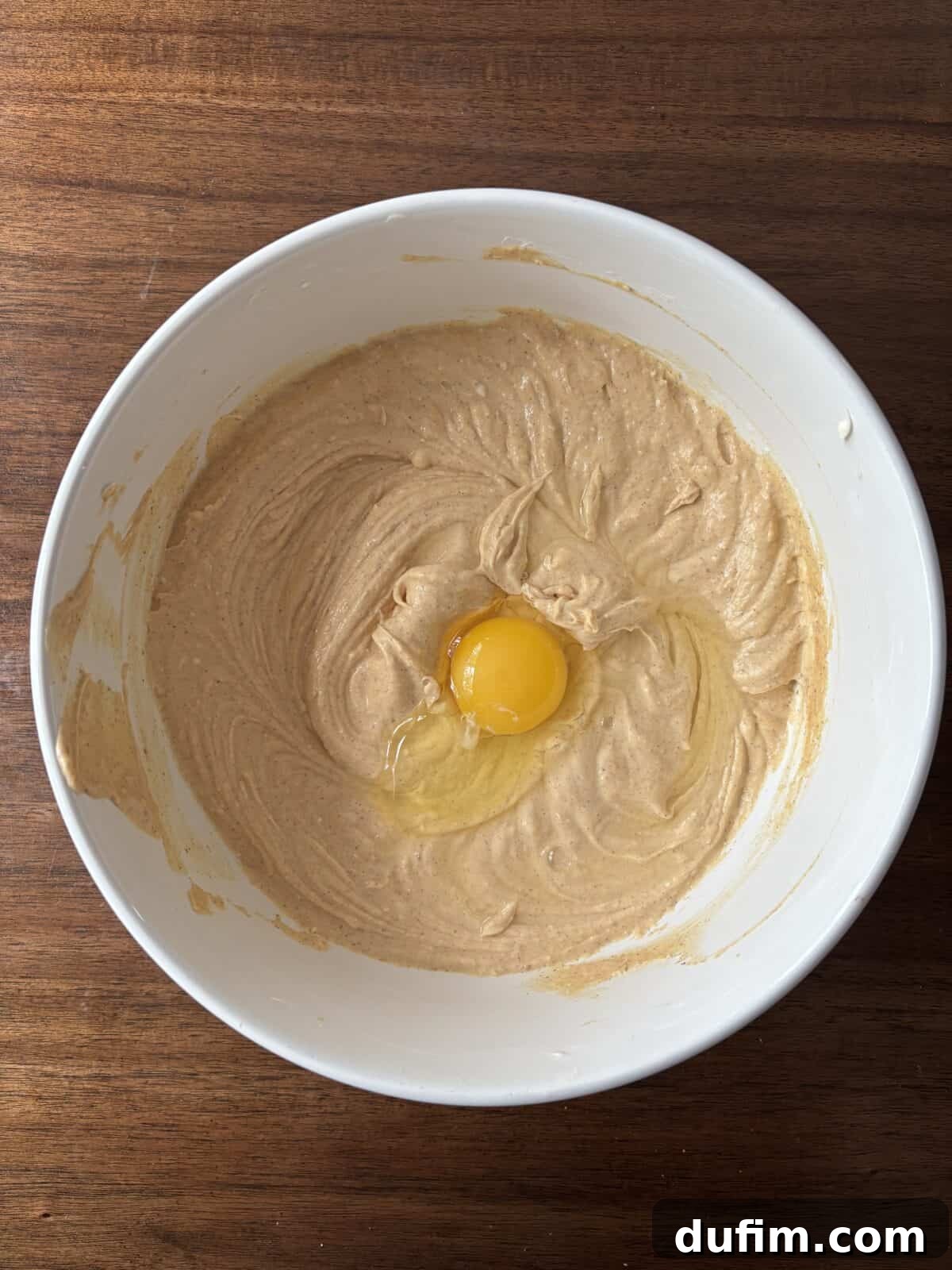A white mixing bowl with creamy light brown batter and a raw egg on top sits on a dark wooden surface.