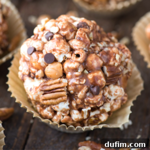 A close-up of a turtle popcorn ball with chocolate drizzle, pecans, chocolate chips, and caramel pieces, sitting in a gold paper cupcake liner on a wooden surface.