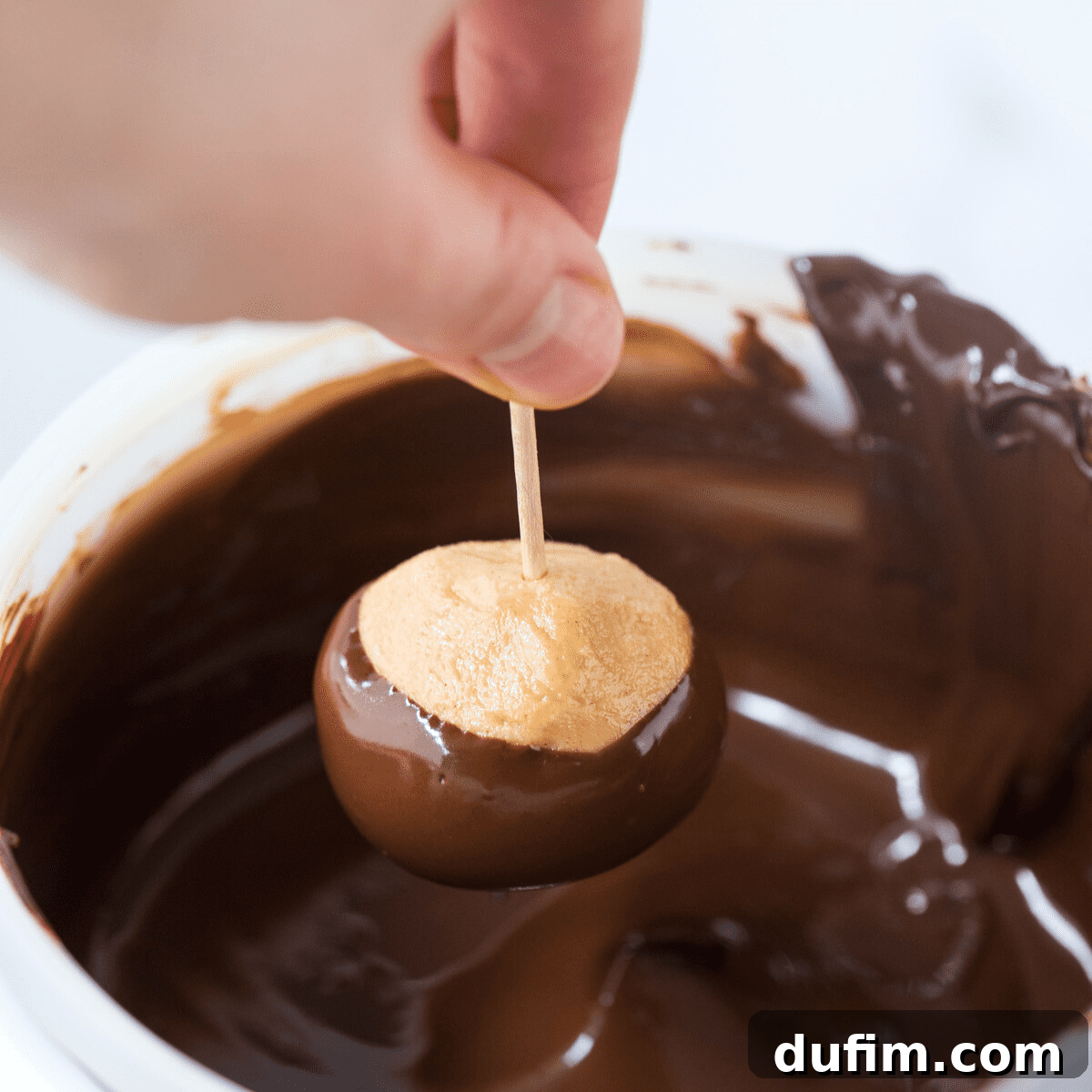 Sweet & Salty Homemade Peanut Butter Buckeye Bites 7 A hand expertly dipping a peanut butter ball into melted chocolate using a toothpick, creating the classic 'buckeye eye'.